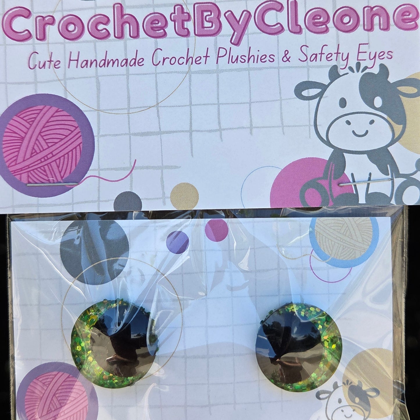A pair of handmade crochet plantain eyes in their original packaging, displayed on a rustic wooden surface. The packaging features a white card with pink, purple, and gray yarn graphics and a cartoon cow illustration.