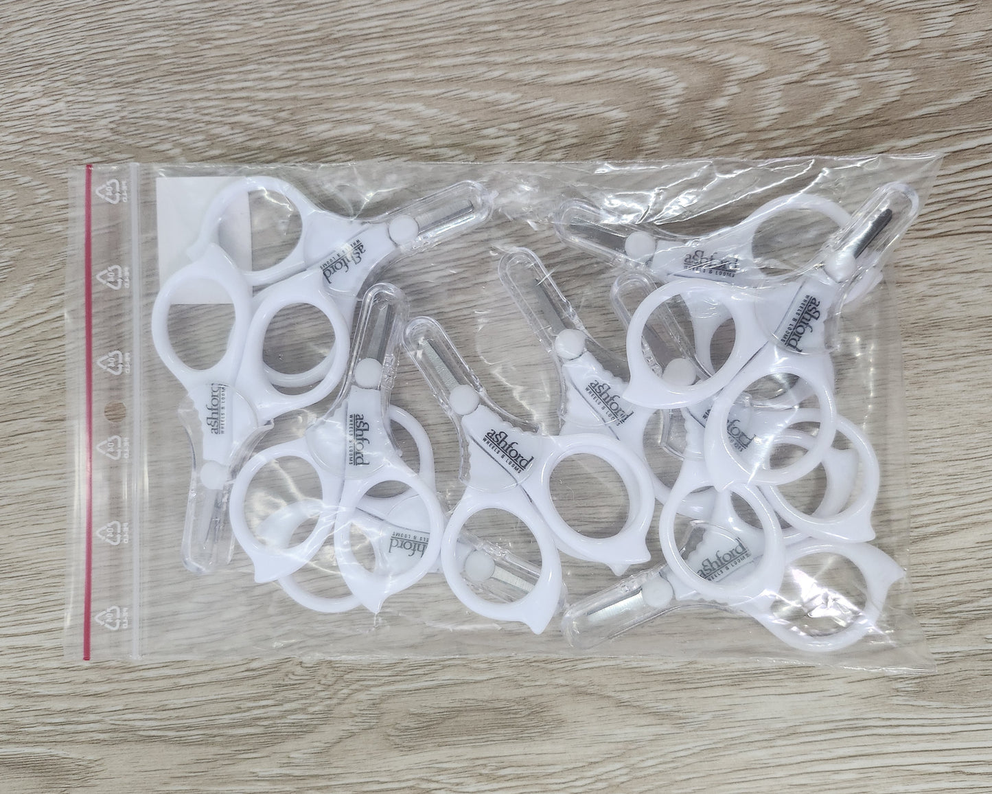 Set of white Ashford Little scissors in a clear plastic bag on a wooden surface