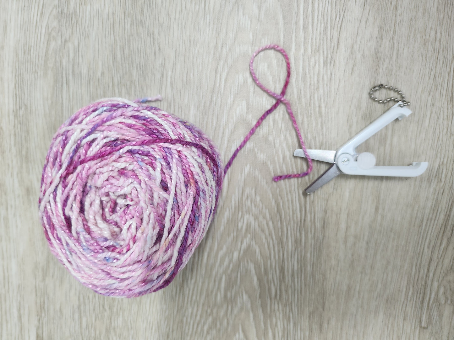 Ball of pink yarn with a string and white foldable retractable scissors on a wooden surface