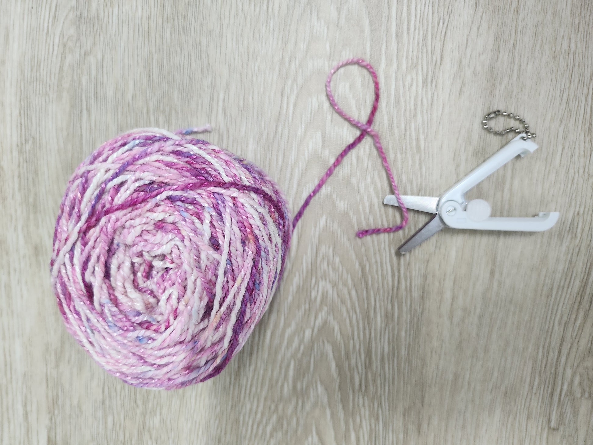 Ball of pink yarn with a string and white foldable retractable scissors on a wooden surface