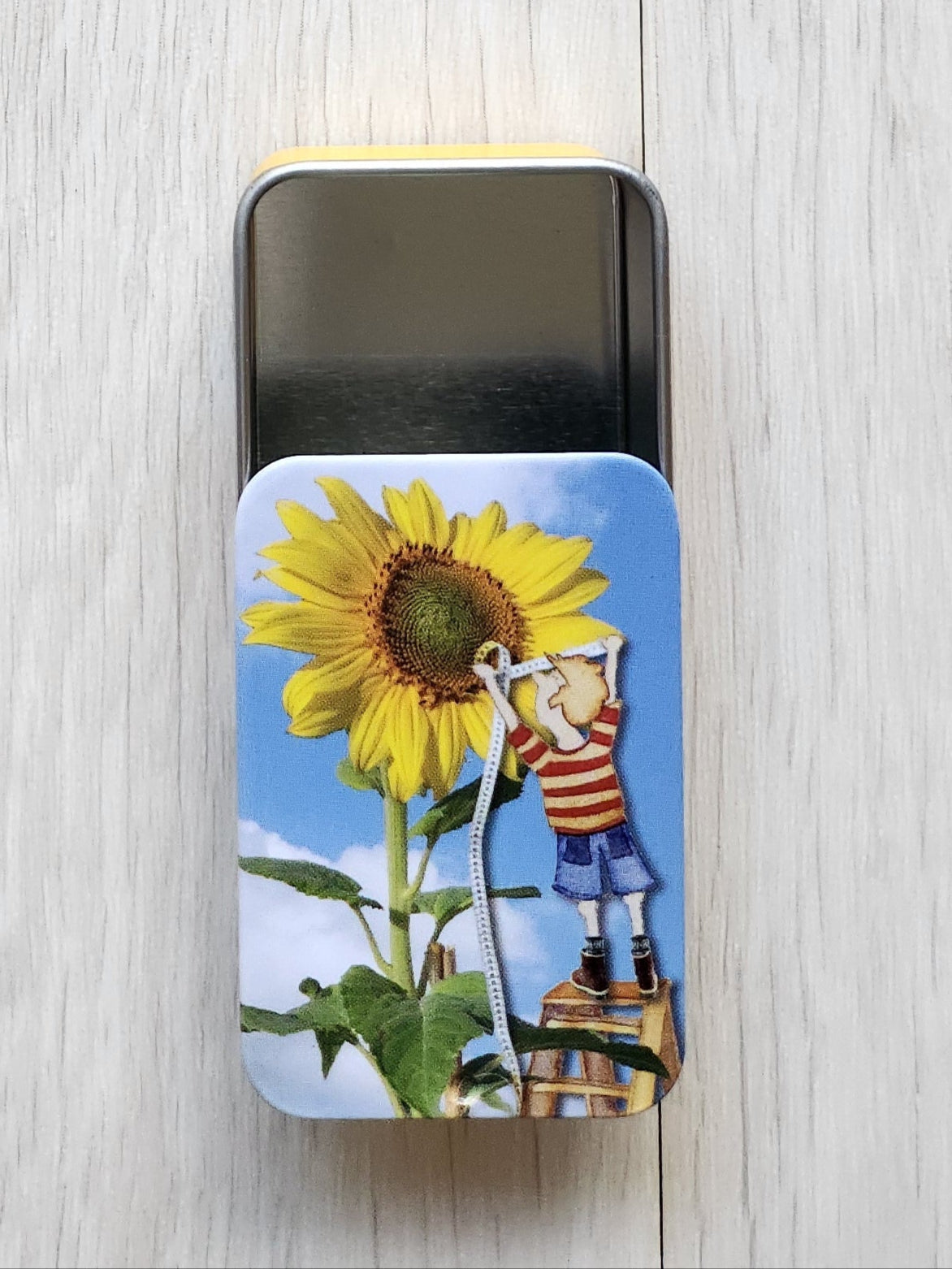 Small tin box with a colorful label featuring a sunflower and two people on a white wooden surface.