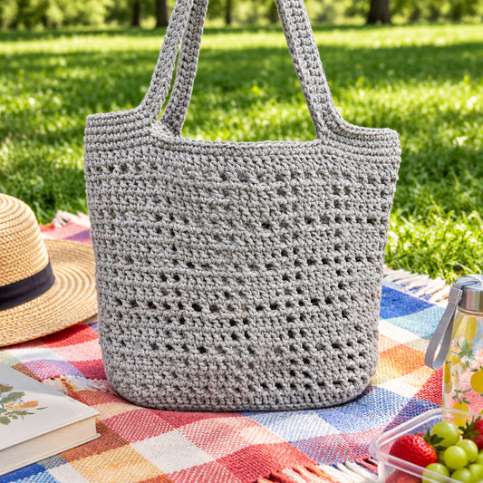 A handmade crochet tote bag in a muted sage green color, hanging against a tan brick wall.