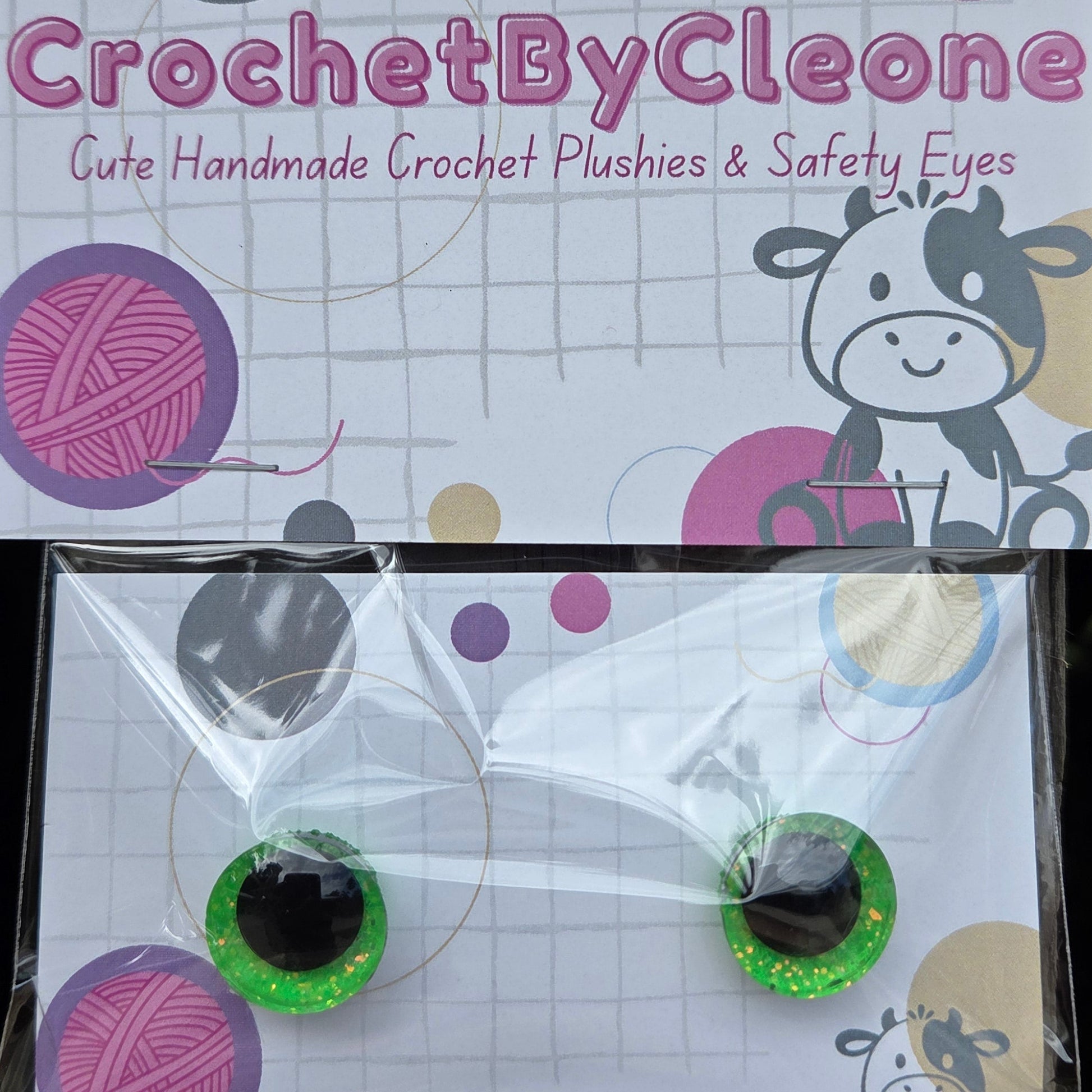 A pair of handmade crochet safety eyes by CrochetByCleone, packaged on a printed card with a cow illustration and yarn ball graphics, hanging against a rustic wooden background.