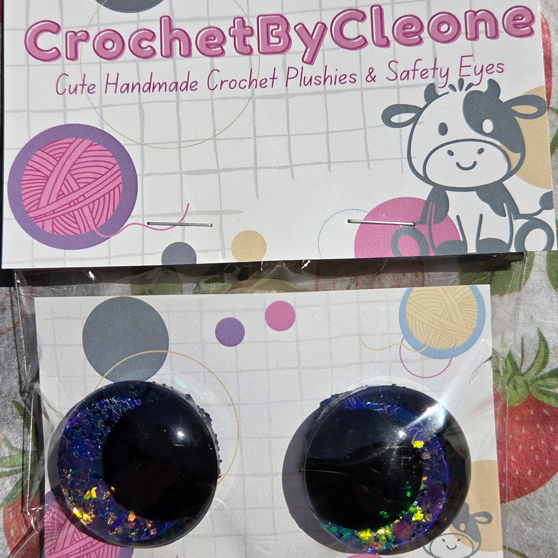 Two packaged crochet hook handles with safety eyes, displayed on a strawberry-patterned fabric background. The left item is in its retail packaging with the brand name 'CrochetByCleone' printed on it, and the right item is in clear plastic wrapping showing the black safety eyes with glittery blue and pink yarn designs.