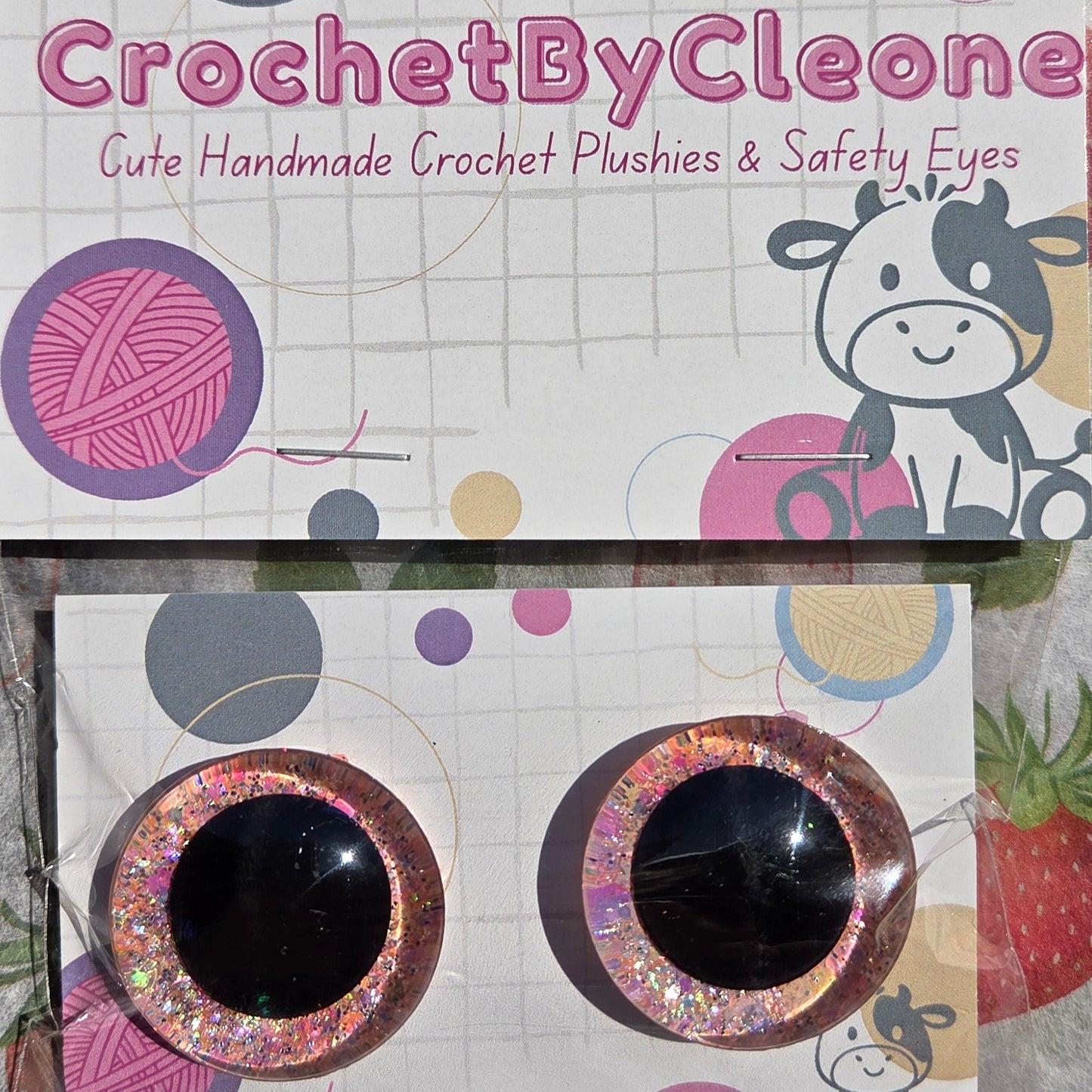 Two handmade crochet patchwork safety eyes with glittery pink frames and black centers, displayed on branded white packaging cards over a strawberry-patterned fabric background.