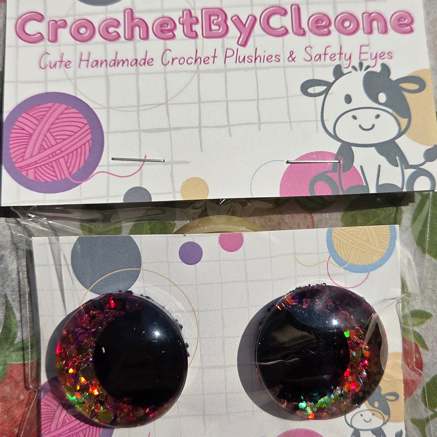 Two handmade crochet plushies in clear plastic packaging on a strawberry-patterned tablecloth. The packaging card reads 'CrochetByCleo' and shows a cow illustration.