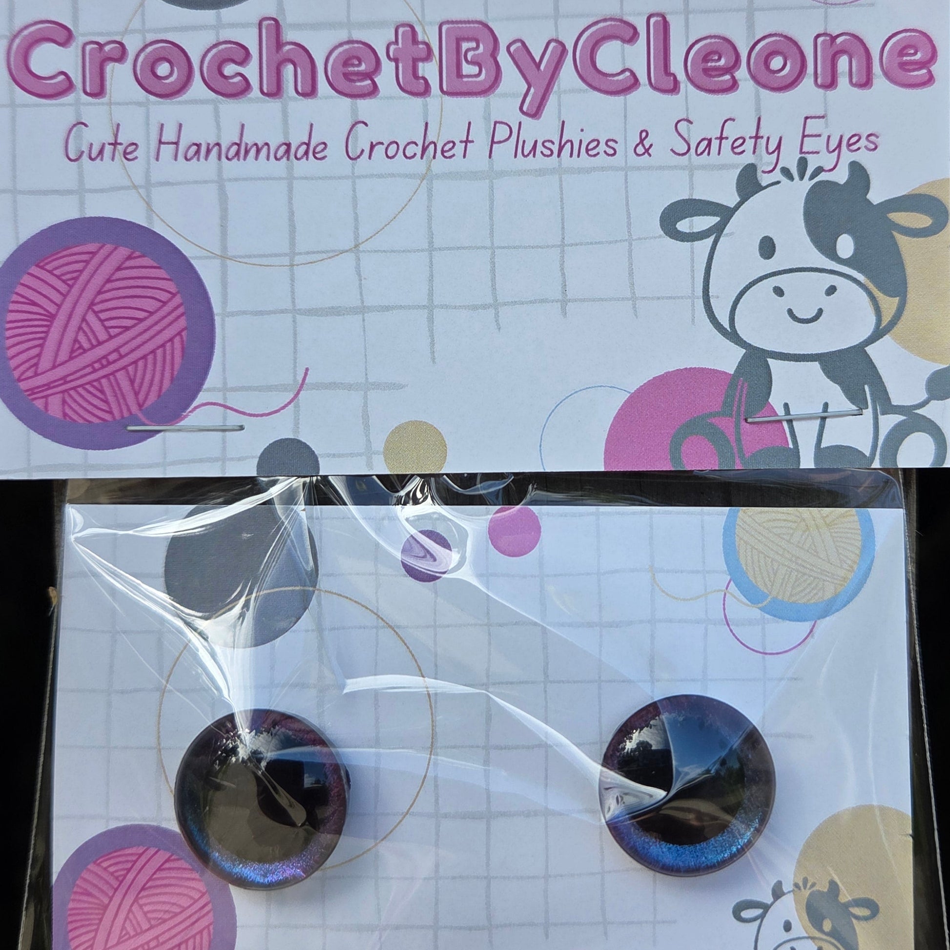Two round crochet plushies with dark blue and purple safety eyes, packaged in a clear plastic sleeve with a printed backing card reading 'CrochetByCleone'.