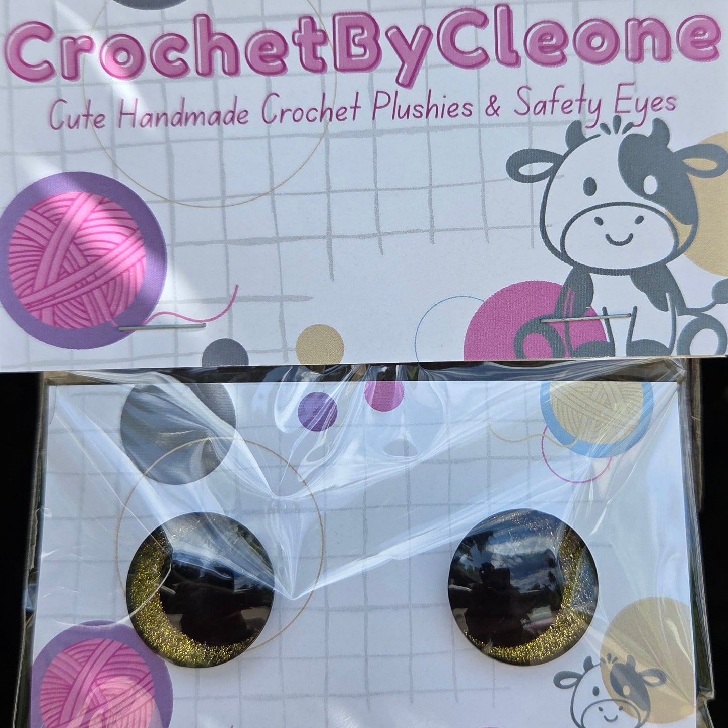 A pair of handmade crochet stud earrings in a clear plastic sleeve, displayed next to their branded cardboard backing card on a rustic wooden surface. The earrings feature a black and gold glittery crochet pattern, and the card is branded 'CrochetByCleone'.