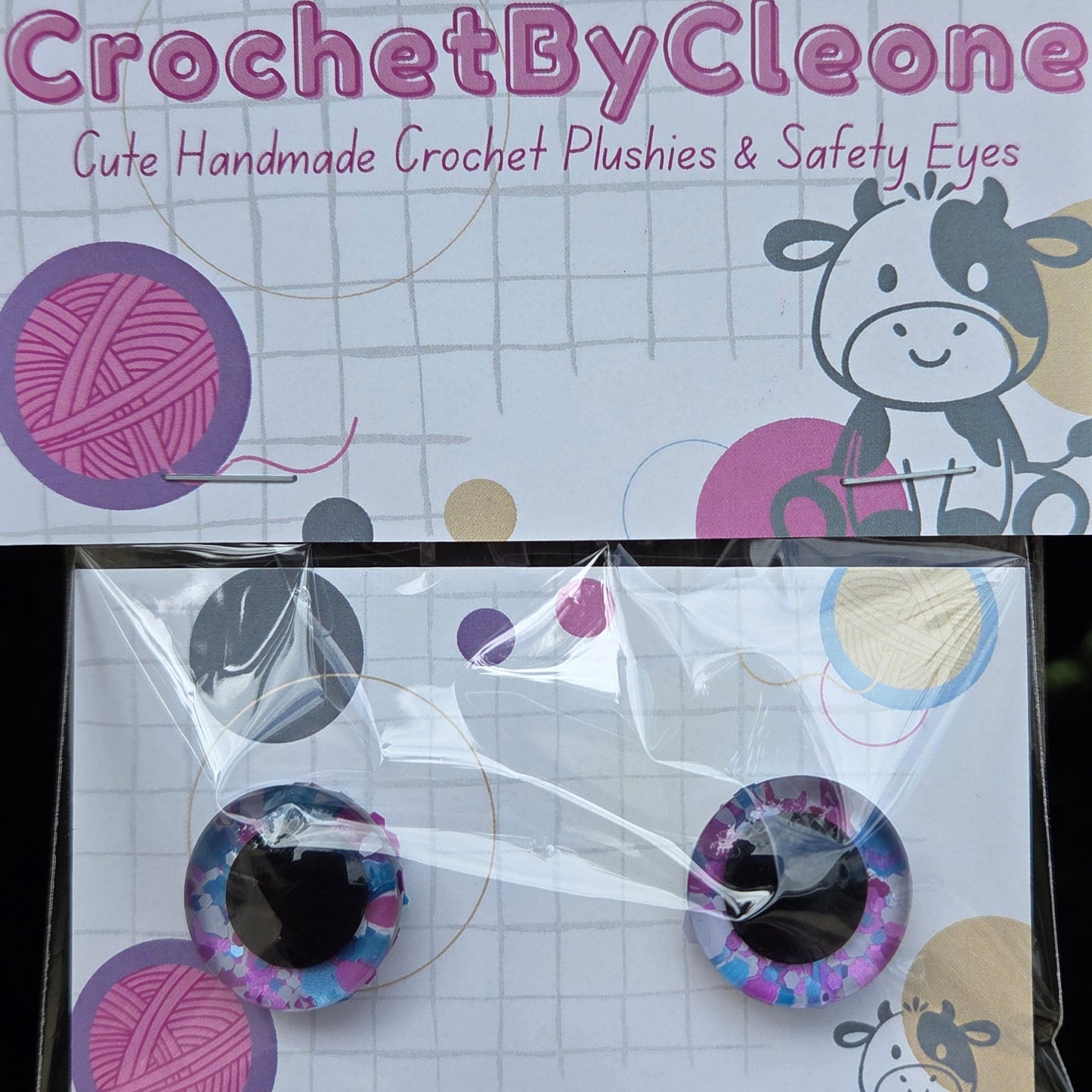 A pair of handmade crochet safety eyes in a purple and blue galaxy pattern, packaged on a white card next to the brand card 'CrochetByCleo'.