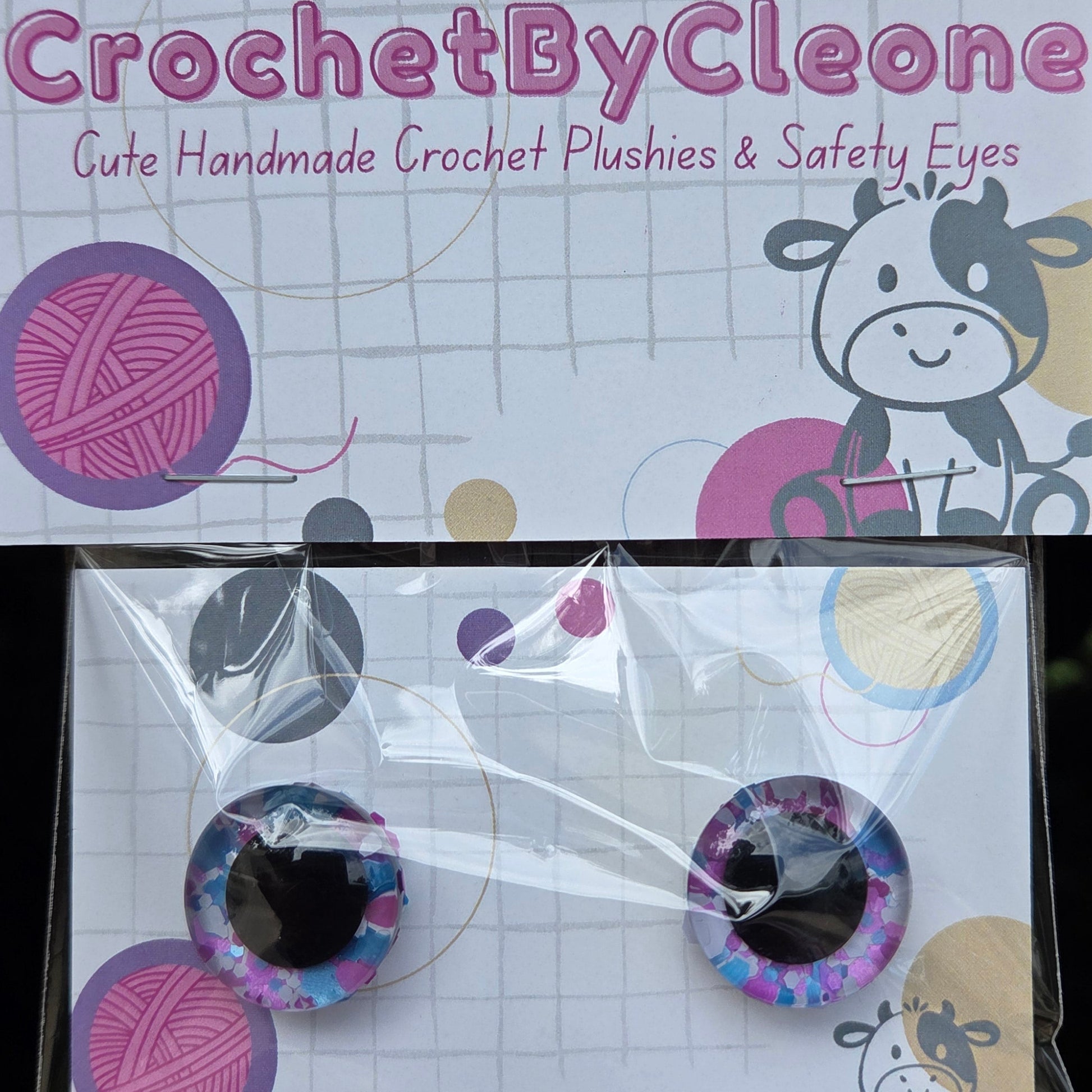 A pair of handmade crochet safety eyes in a purple and blue galaxy pattern, packaged on a white card next to the brand card 'CrochetByCleo'.