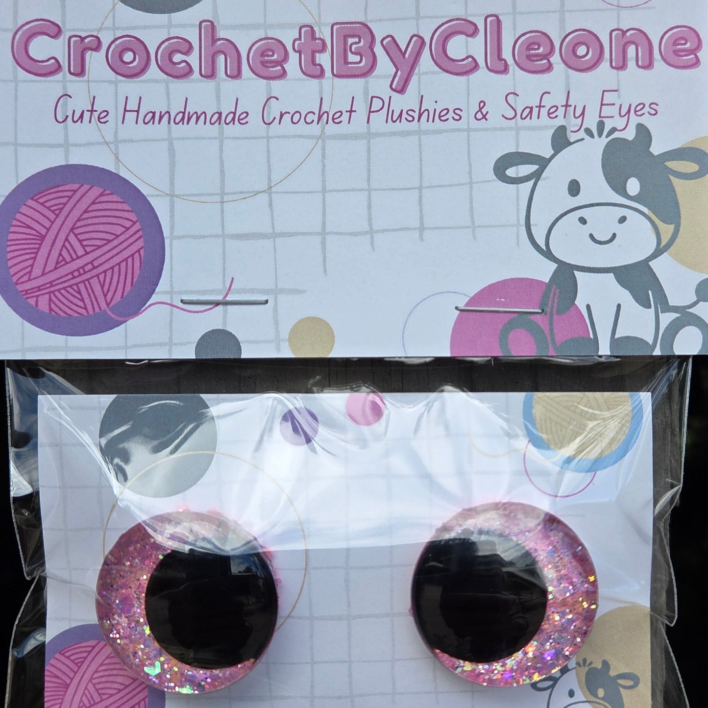 Two round novelty crochet punch holes in glittery pink and silver, displayed on a white backing card next to the product's packaging card on a rustic wooden surface.