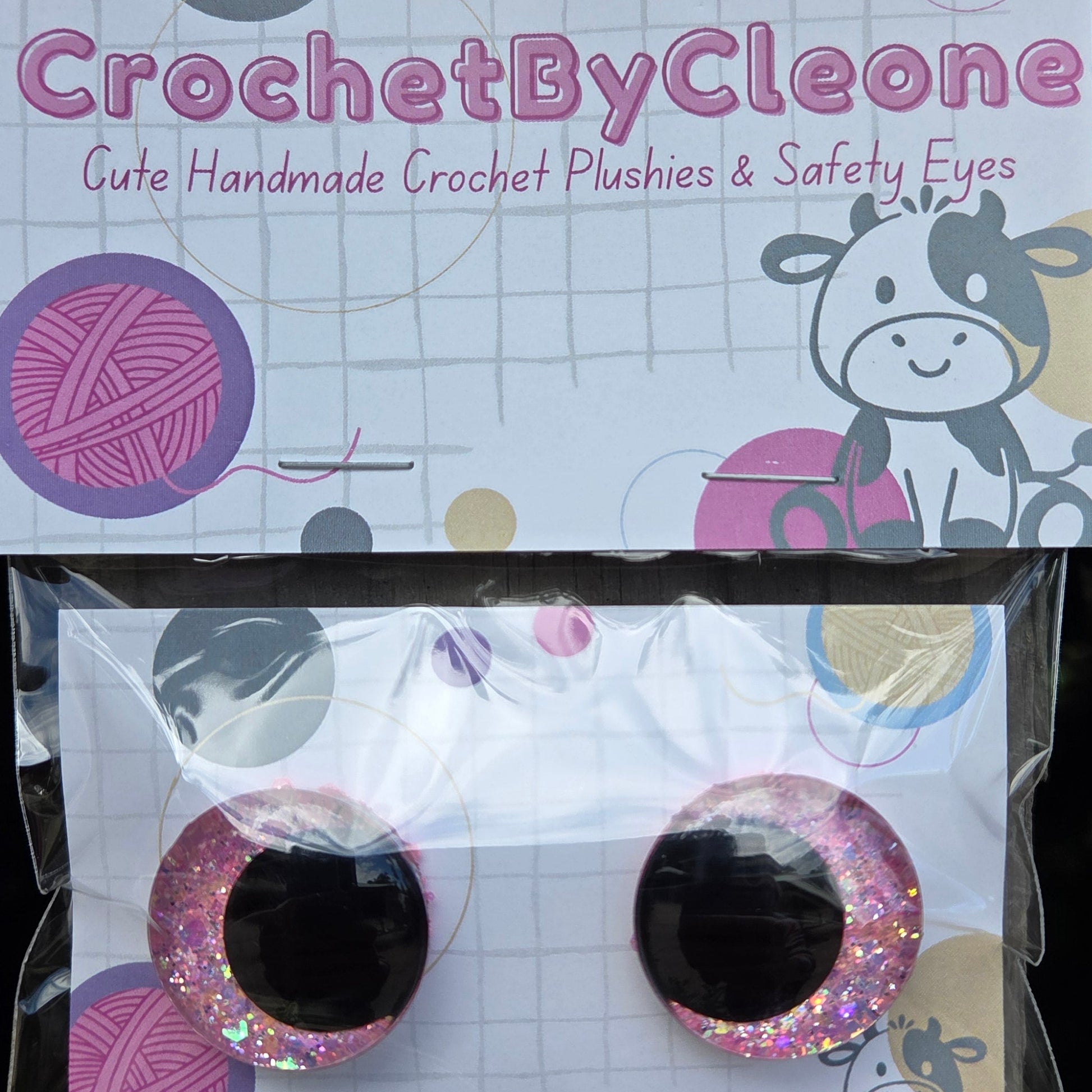 Two round novelty crochet punch holes in glittery pink and silver, displayed on a white backing card next to the product's packaging card on a rustic wooden surface.