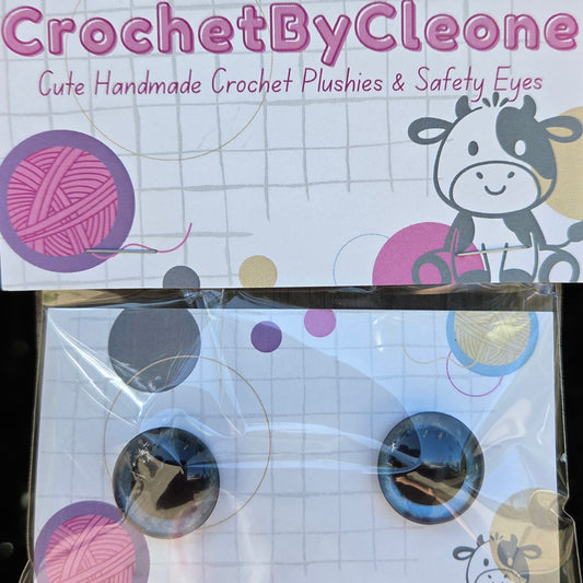 A pair of large novelty crochet stitches inlaid with black, white, and gold glitter, mounted on a white card with 'CrochetByCleo' branding, displayed on a rustic wooden surface.
