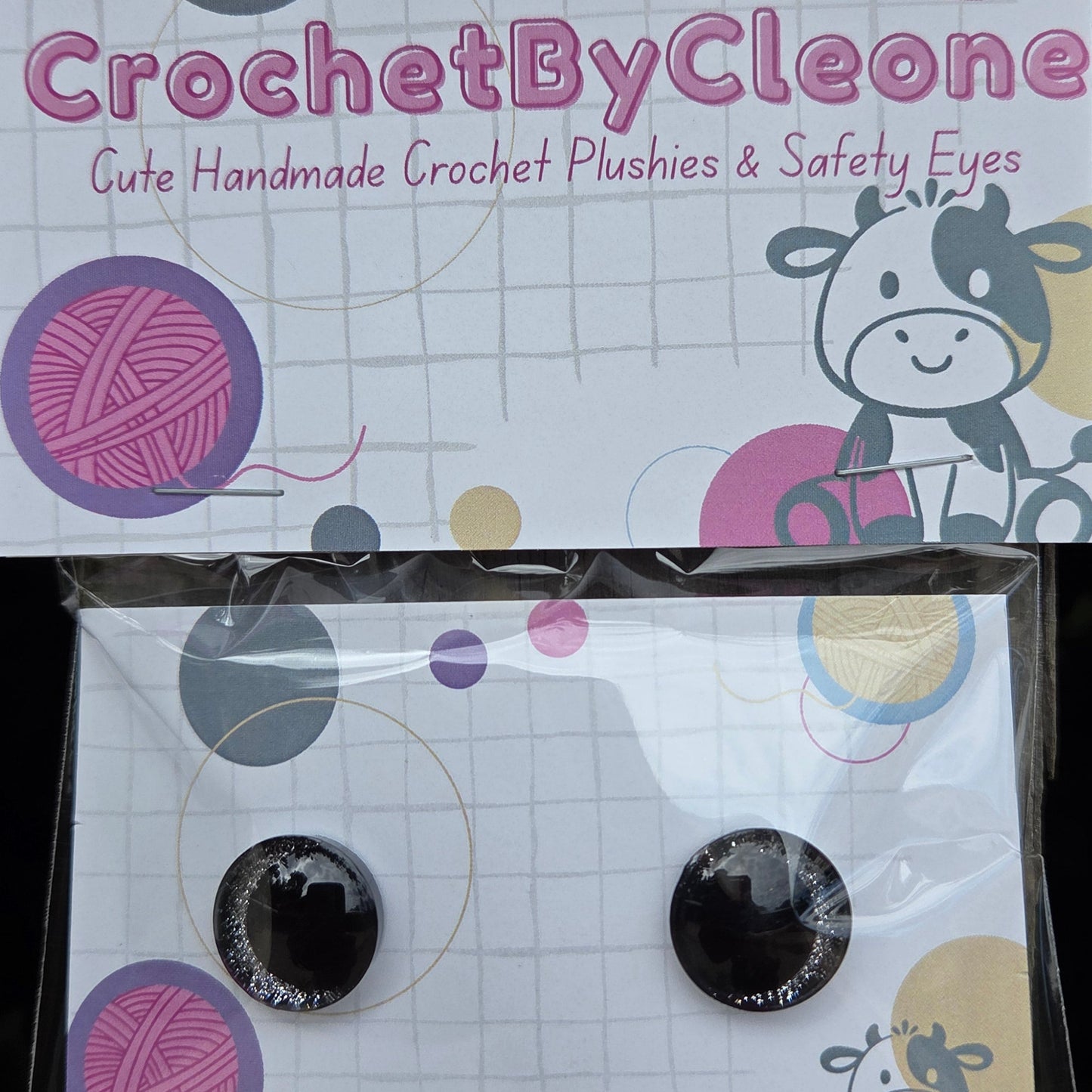 A pair of handmade crochet eyelashes and safety eyes in a clear plastic sleeve, displayed next to its branded cardboard backing card on a rustic wooden surface.