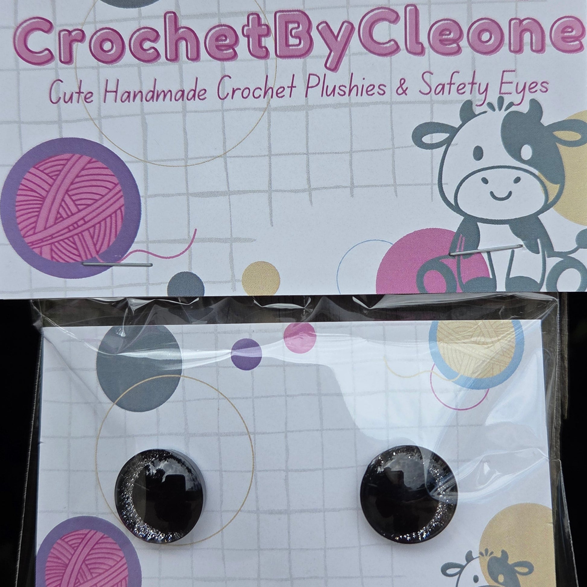A pair of handmade crochet eyelashes and safety eyes in a clear plastic sleeve, displayed next to its branded cardboard backing card on a rustic wooden surface.