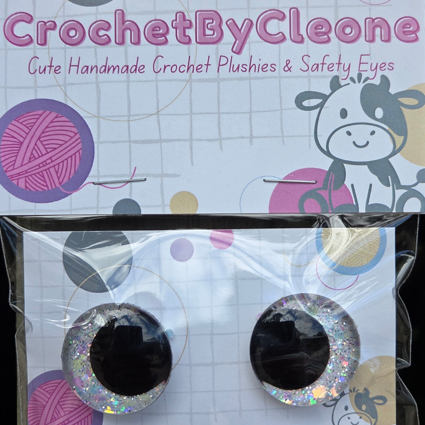 A pair of handmade crochet plastic and safety eyes with glittery silver borders, displayed on a branded card next to its packaging. The packaging features the brand name 'CrochetByCleone' and illustrations of yarn balls and a cow.