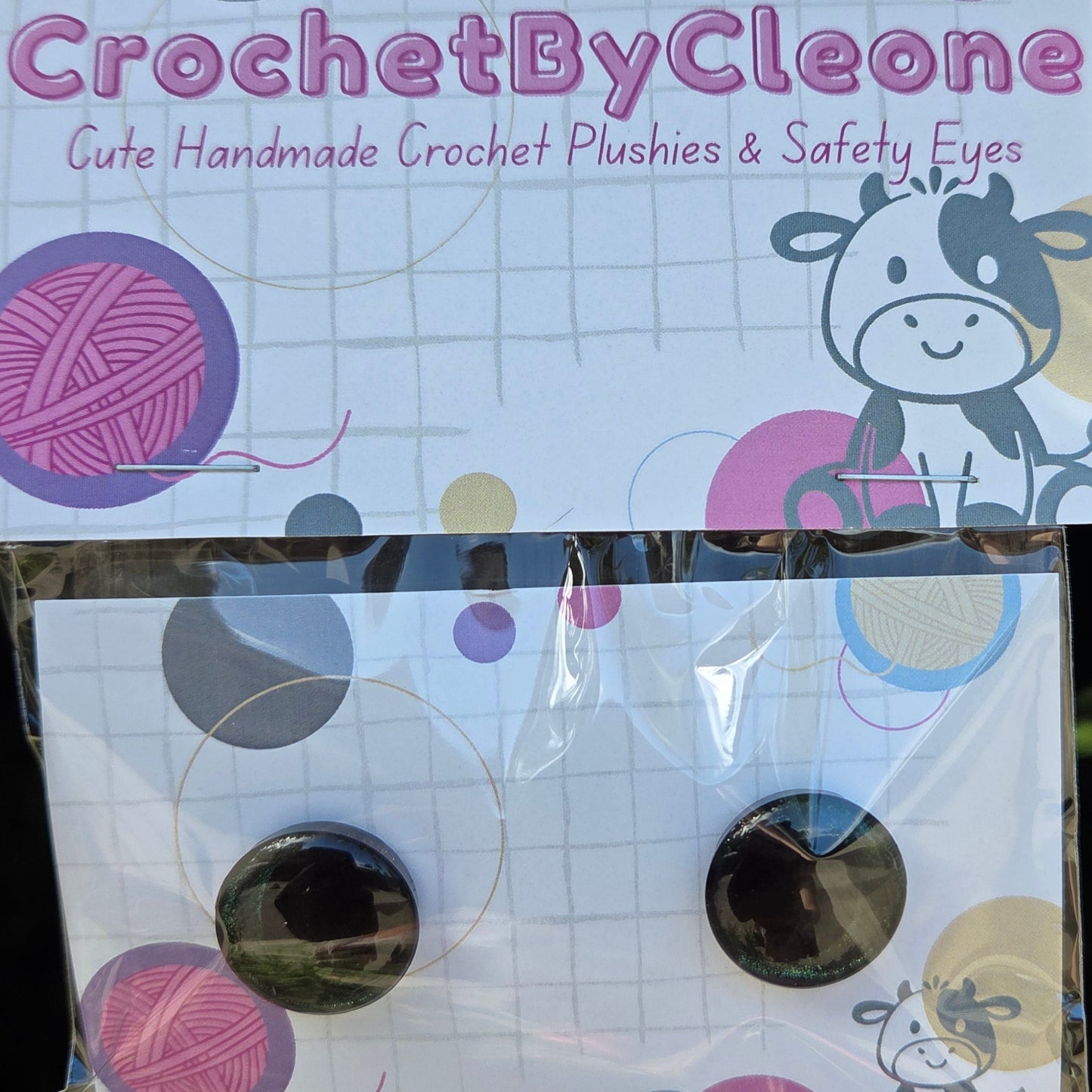 A pair of handmade crochet safety eyes in their original packaging, displayed on a rustic wooden surface. The packaging features a white card with pink and purple yarn illustrations and the brand name 'CrochetByCleone'.
