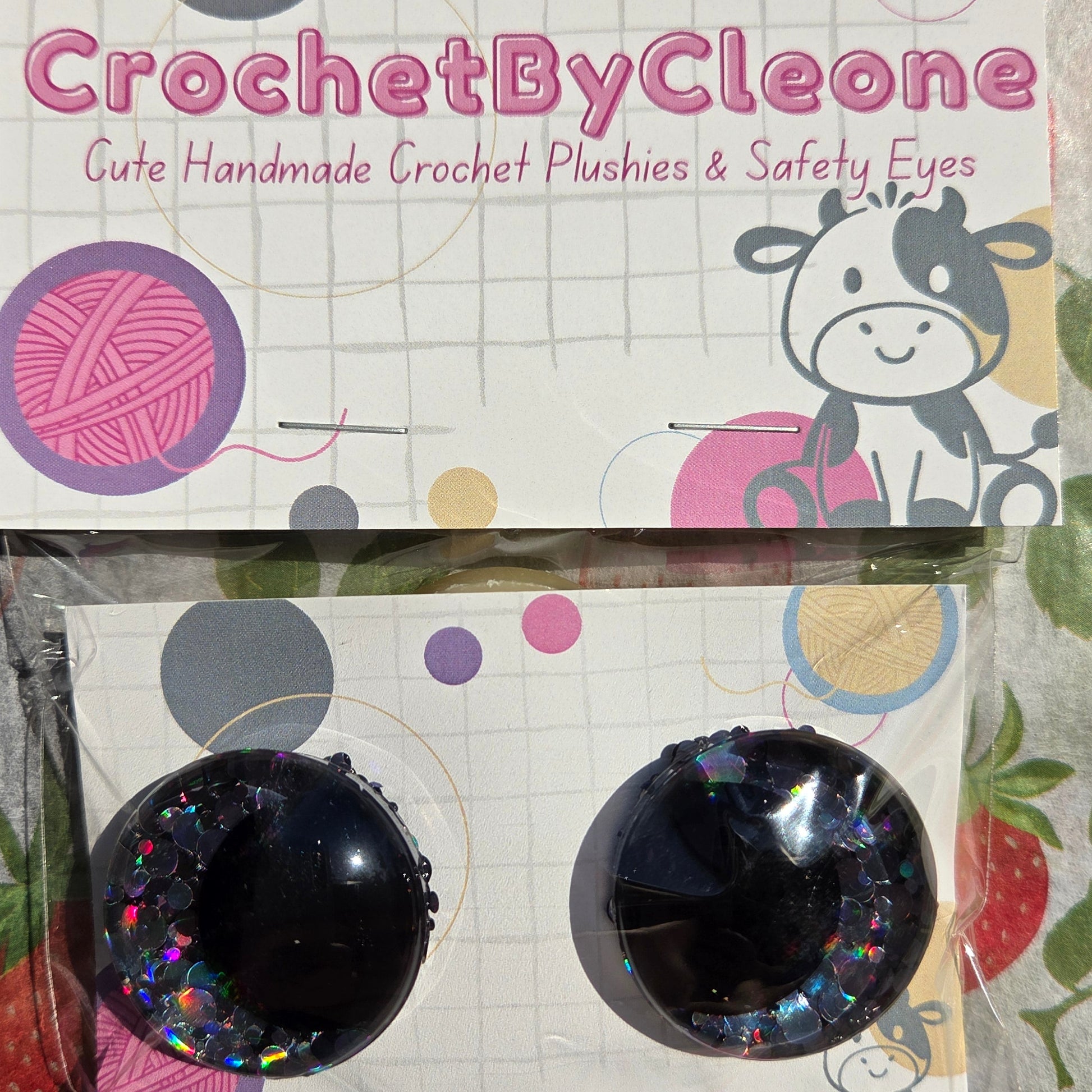 Two round black crochet crochet hook eyes with iridescent glitter accents, displayed on a branded card next to its packaging on a strawberry-patterned fabric background.