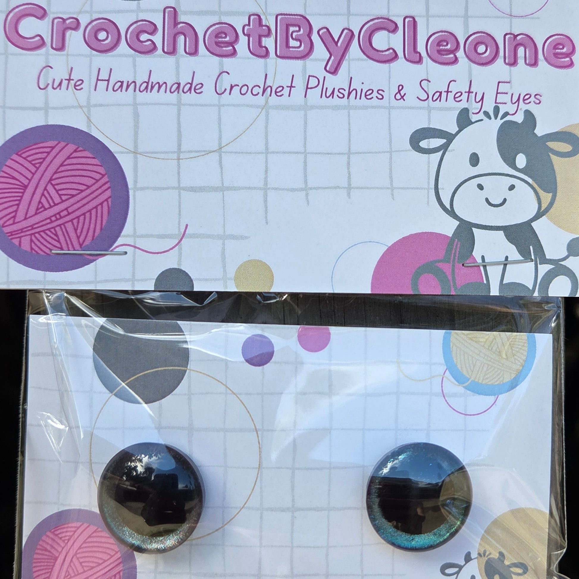 A pair of handmade crochet safety eyes displayed on a branded backing card. The card reads 'CrochetByCleone' and shows colorful yarn ball graphics and a cow illustration.