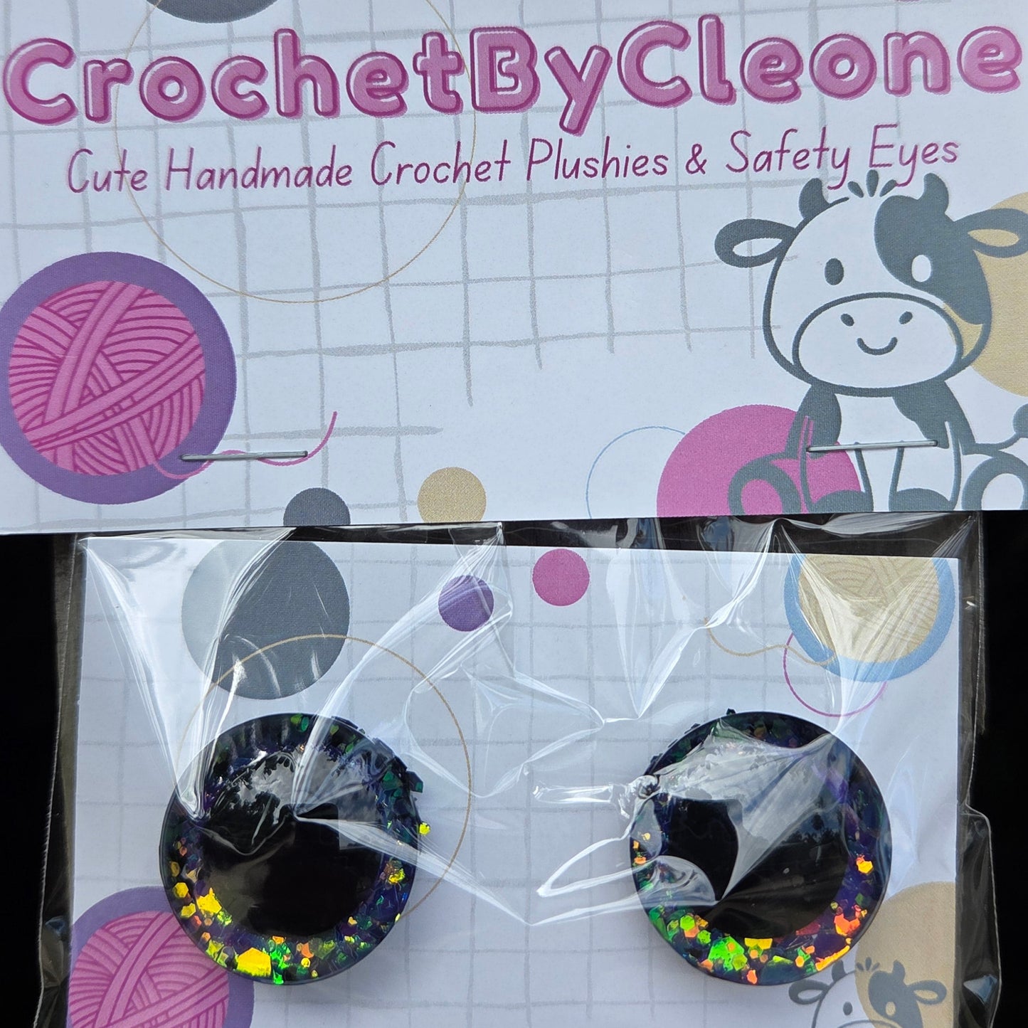 A pair of handmade crochet safety eyes by CrochetByCleo, packaged on a branded card and clear plastic sleeve, displayed on a rustic wooden surface.