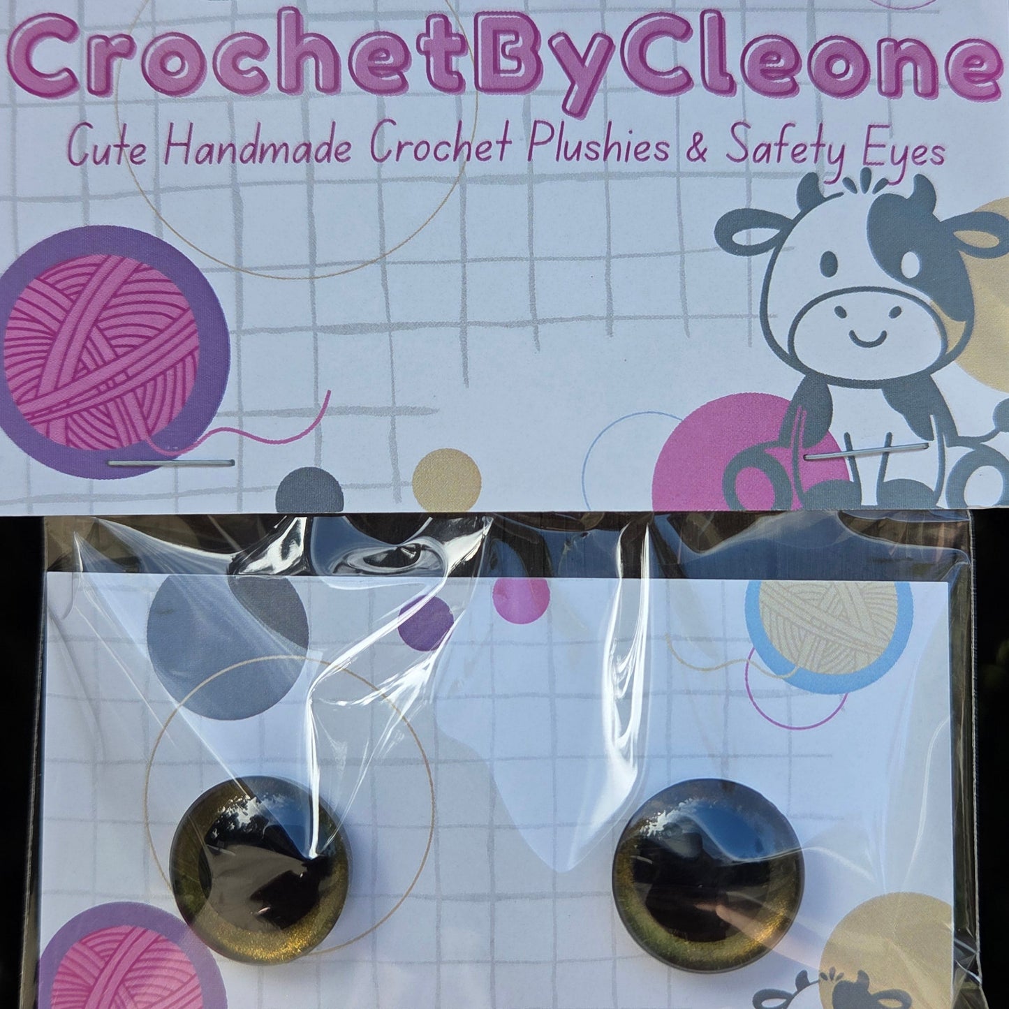 A pair of round cat-eye style crochet hook plungers in a clear plastic sleeve, displayed next to its branded packaging card. The packaging features a white background with a grid pattern, cartoon cow illustrations, and colorful yarn ball graphics.