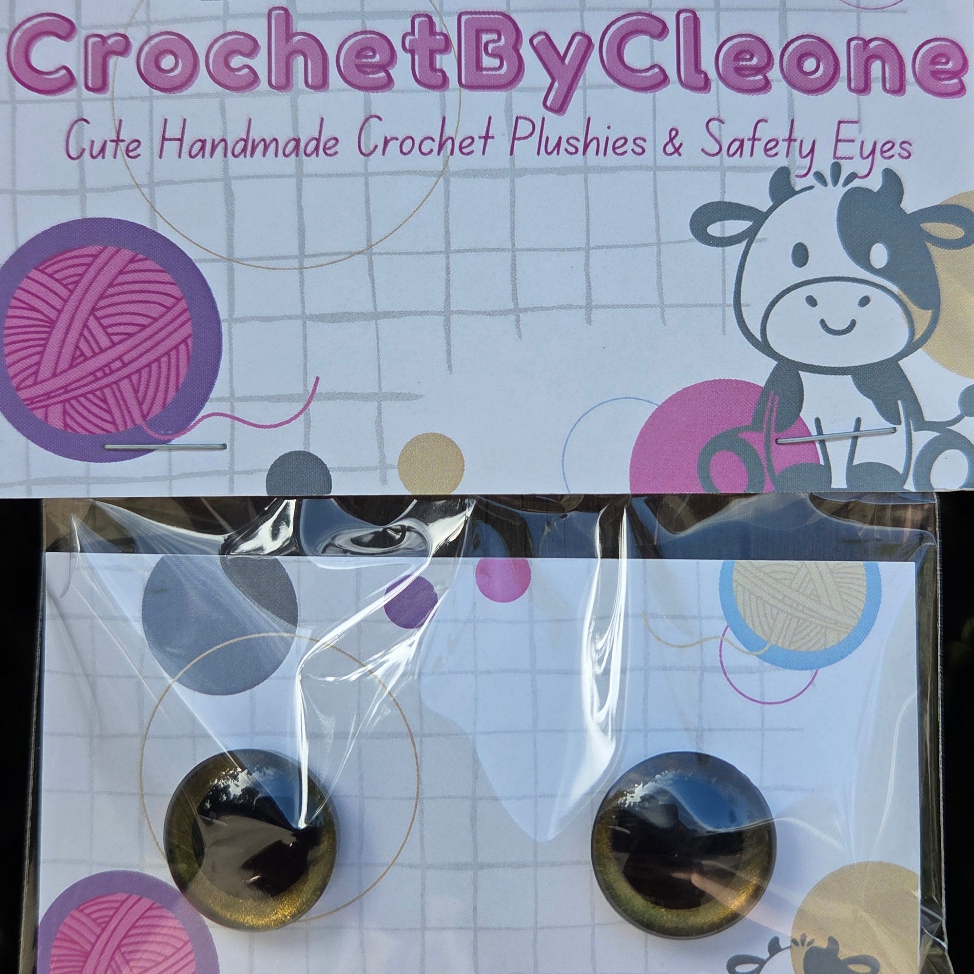 A pair of round cat-eye style crochet hook plungers in a clear plastic sleeve, displayed next to its branded packaging card. The packaging features a white background with a grid pattern, cartoon cow illustrations, and colorful yarn ball graphics.