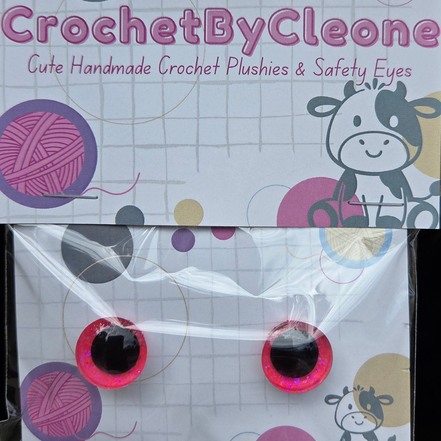 A pair of handmade crochet crochet eyes by CrochetByCleo, packaged on a branded white card with colorful yarn and cow illustrations, resting on a rustic wooden surface.