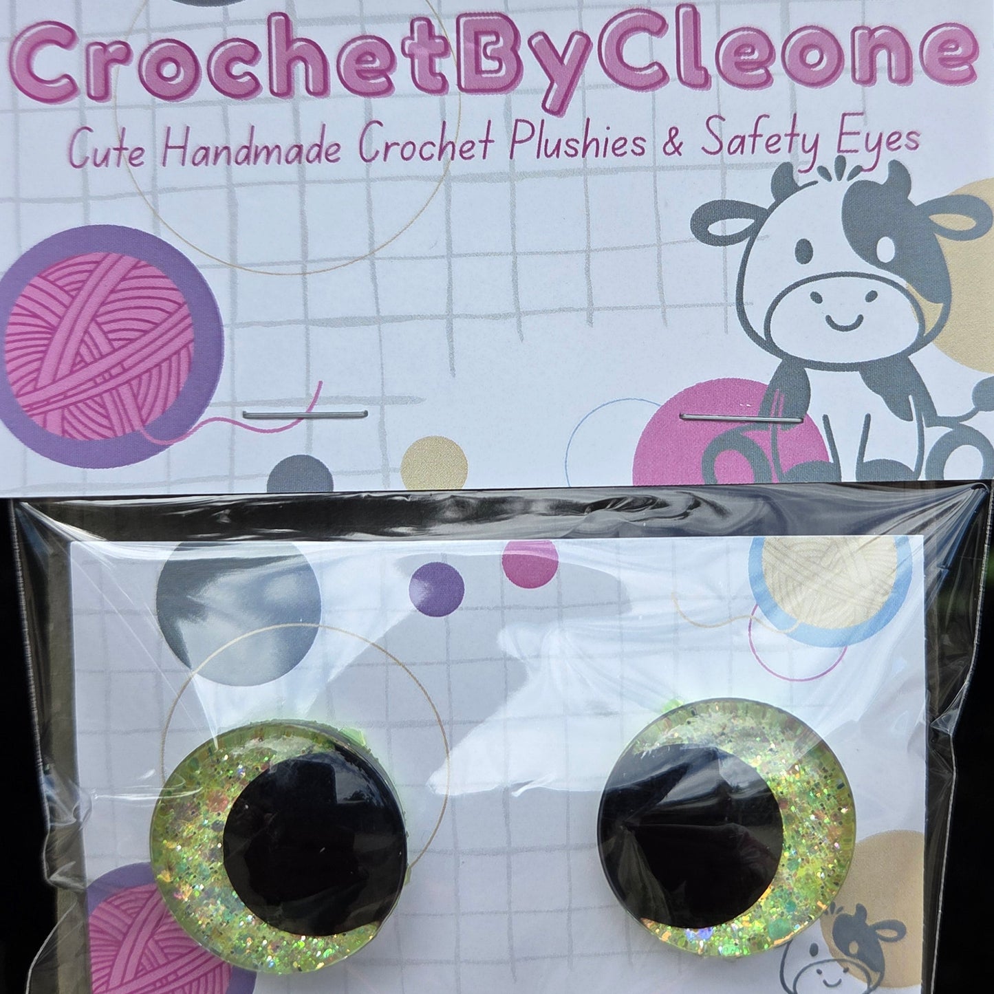 A pair of handmade crochet crochet plushies and safety eyes by CrochetByCleo, shown with their branded packaging card on a rustic wooden surface.
