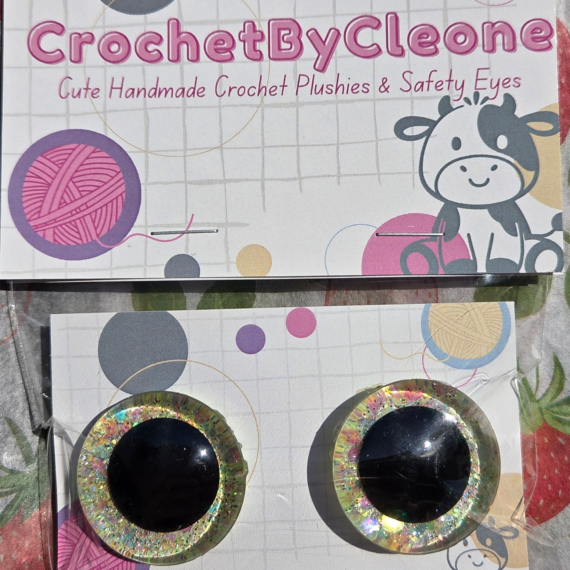 Two handmade crochet punch holes with glittery circular frames and black centers, displayed on branded cards over a strawberry-patterned fabric background.