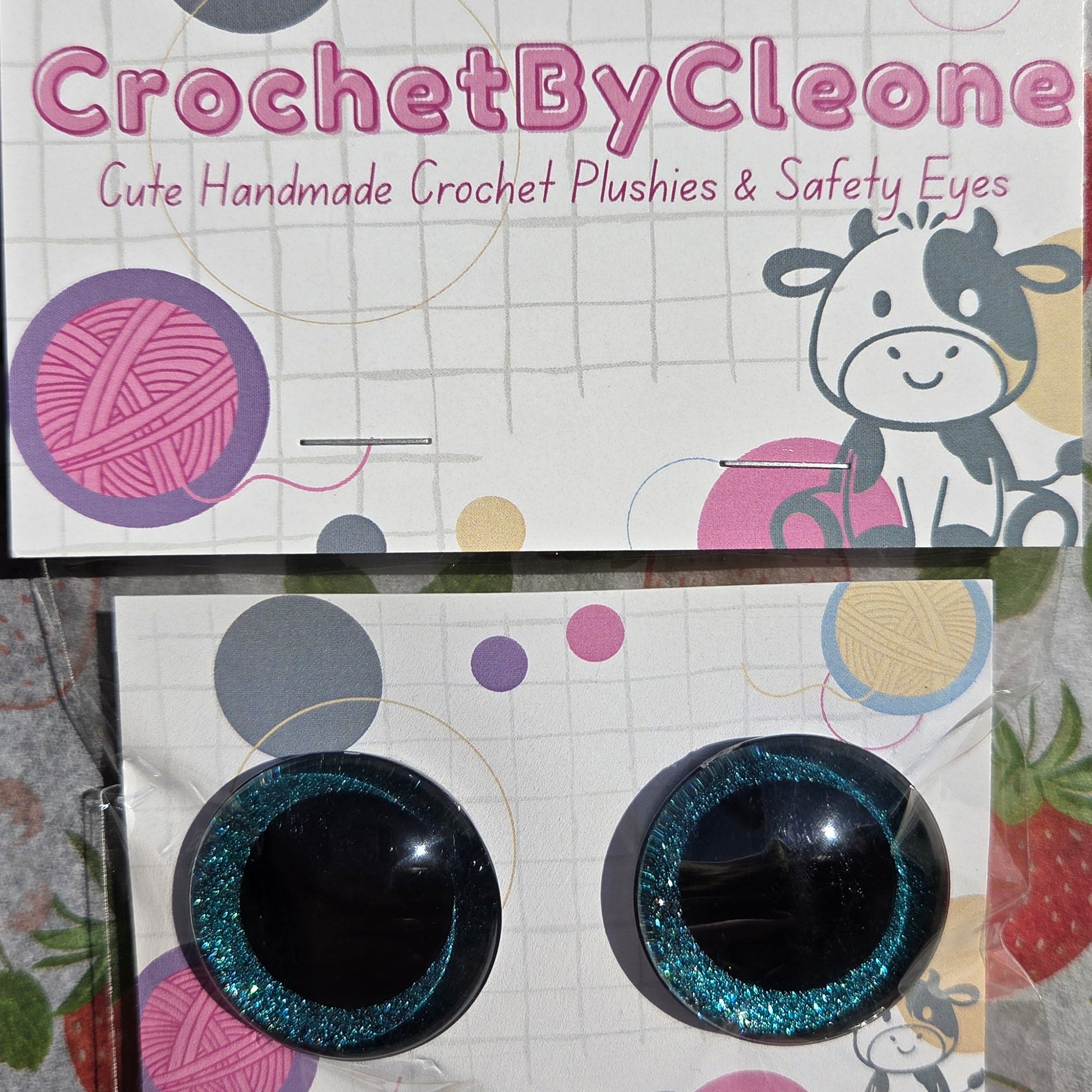 A pair of round black crochet safety eyes with glittery teal-blue rims, displayed on a branded white backing card on a strawberry-patterned fabric surface.