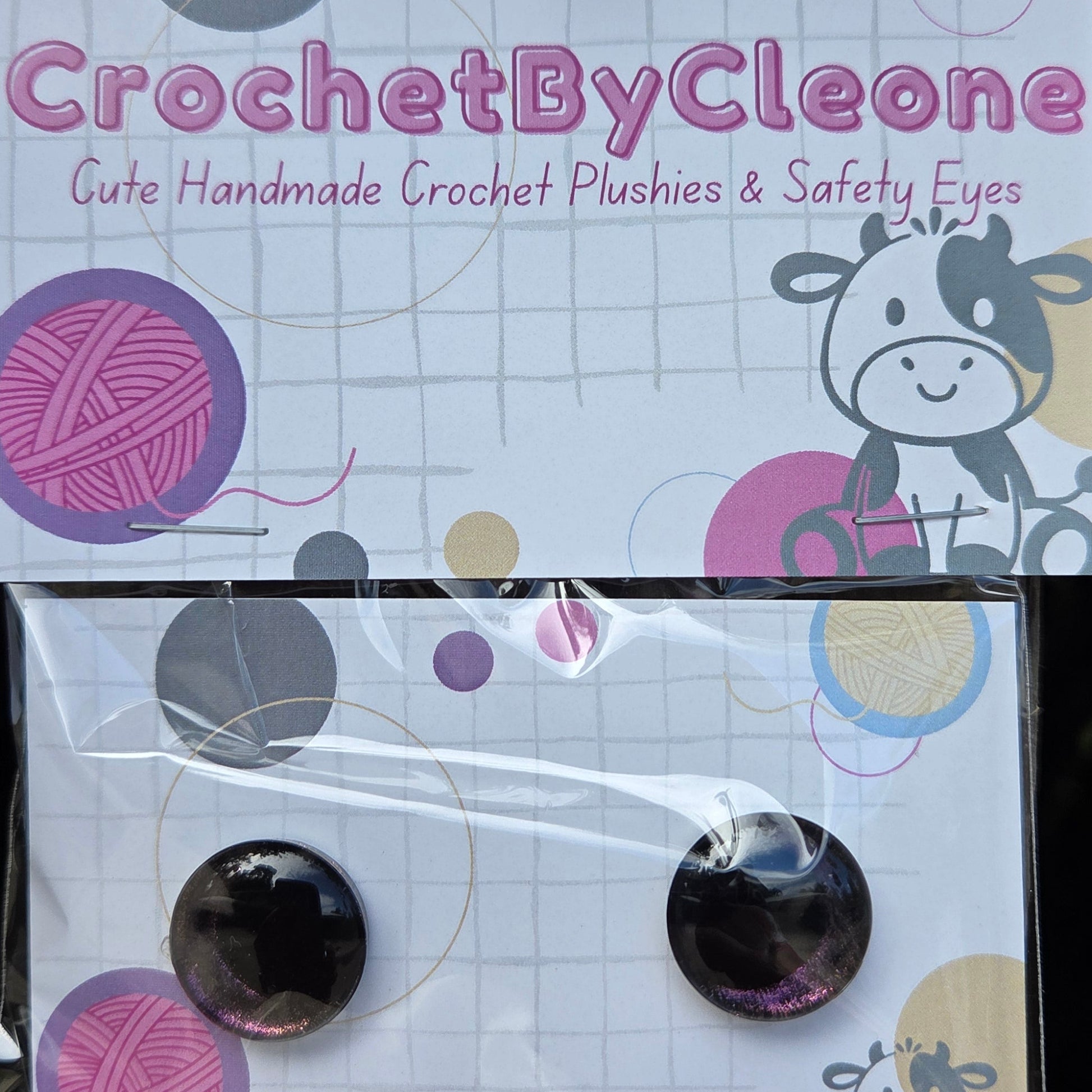 A pair of handmade crochet safety eyes displayed on a branded backing card. The round black safety eyes are attached to a white card printed with pink yarn illustrations, a cow graphic, and the brand name 'CrochetByCleone'.