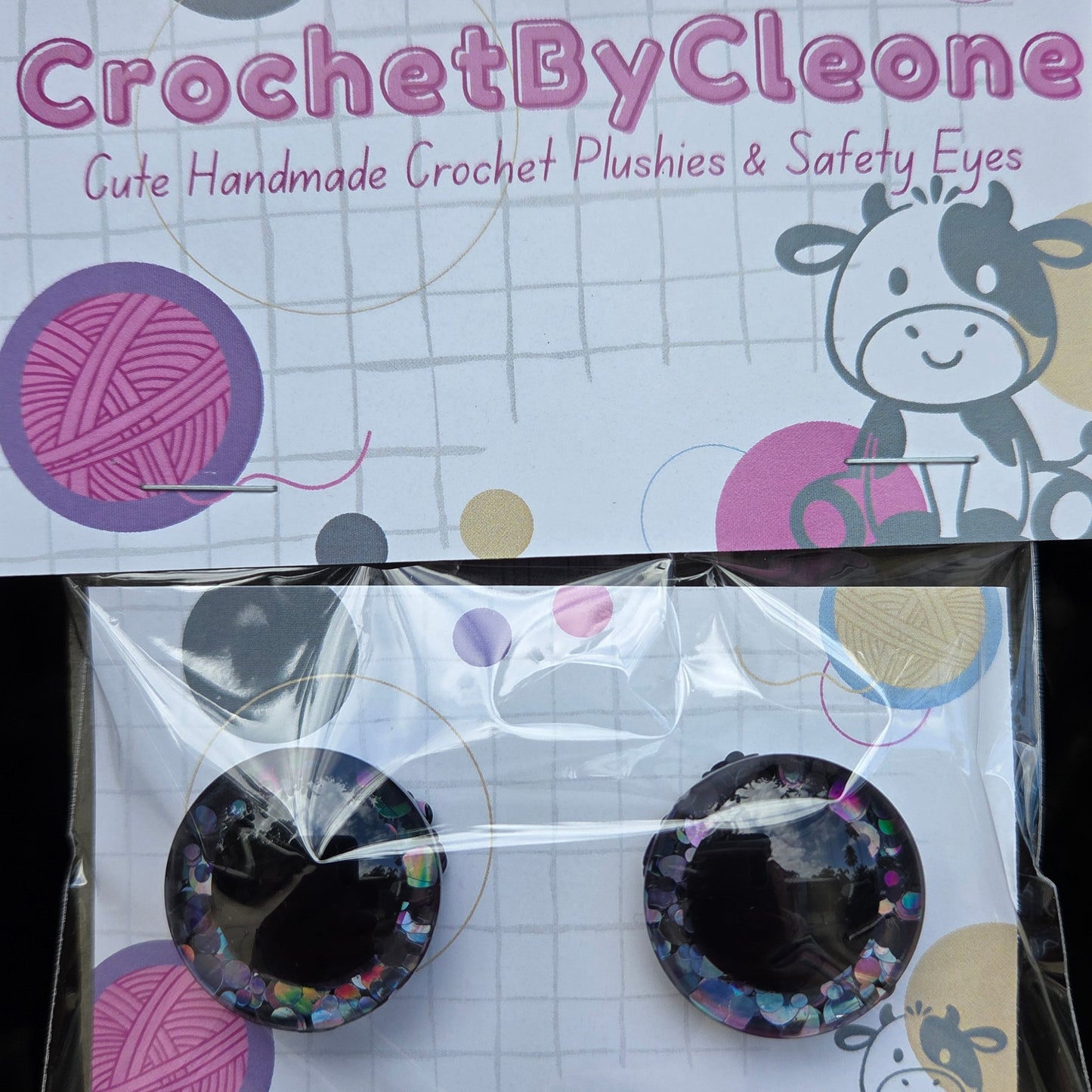 A pair of handmade crochet safety eyes with glittery multicolor accents, packaged on a white card with cartoon cow illustrations and the brand name 'CrochetByCleo' printed on the sleeve.
