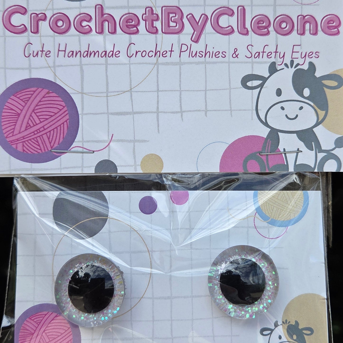 Two packaged novelty crochet eyes displayed on branded cards on a rustic wooden surface. The cards read 'CrochetByCleo' and show the product as cute handmade crochet plushies and safety eyes.