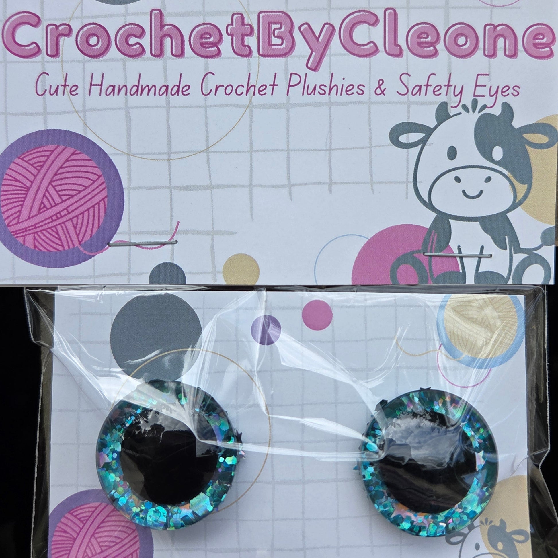 A pair of handmade crochet safety eyes in a clear plastic sleeve, displayed next to their branded packaging card on a rustic wooden surface. The eyes feature glittery teal-blue and black glitter borders with clear plastic centers.