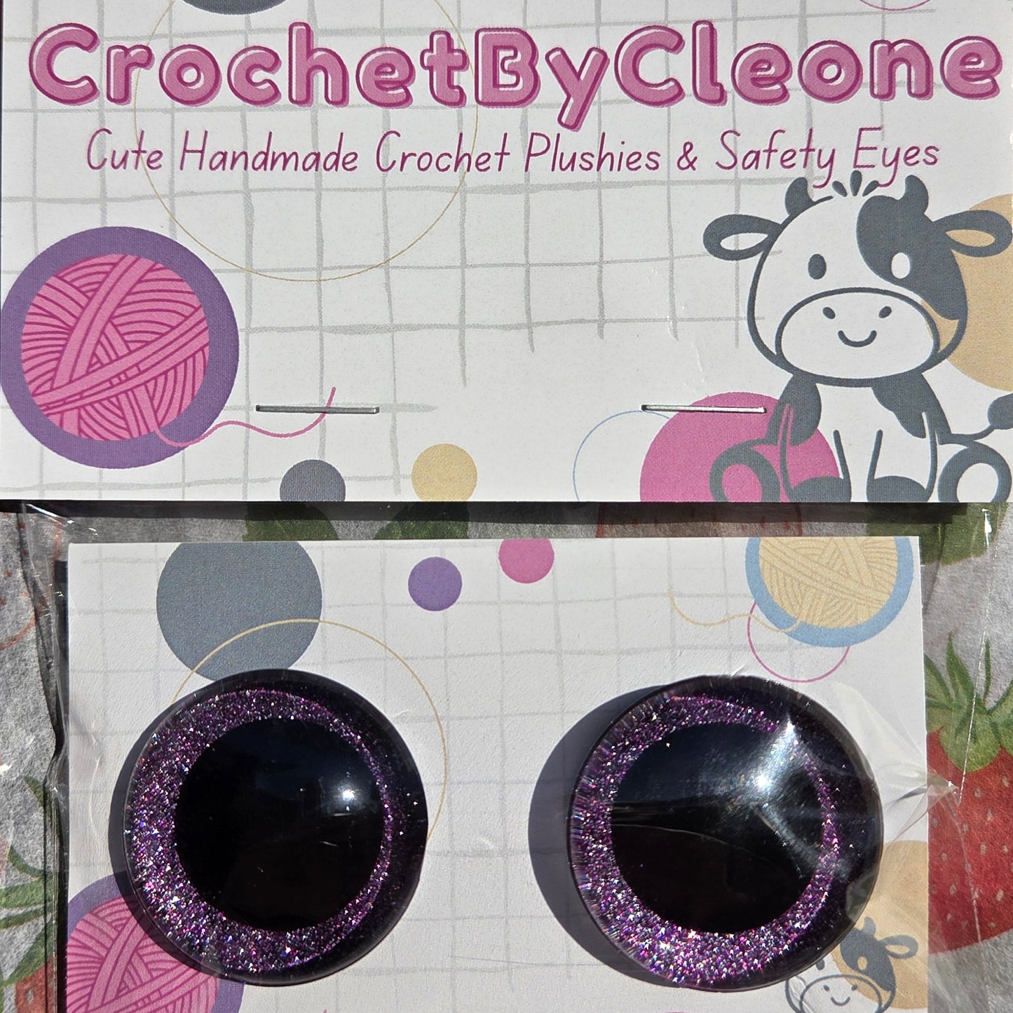 Two round black and glittery purple crochet hooks in their original branded packaging, displayed on a strawberry-patterned surface.