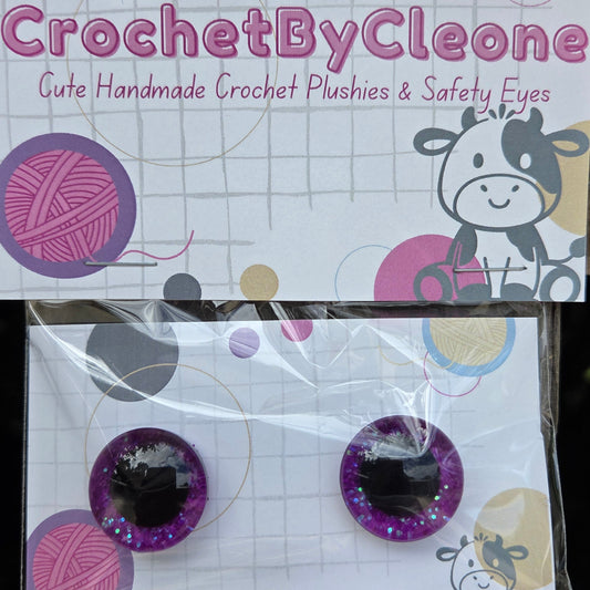 A pair of handmade crochet plushies in purple glitter with black centers, displayed on a branded card next to its packaging on a rustic wooden surface.