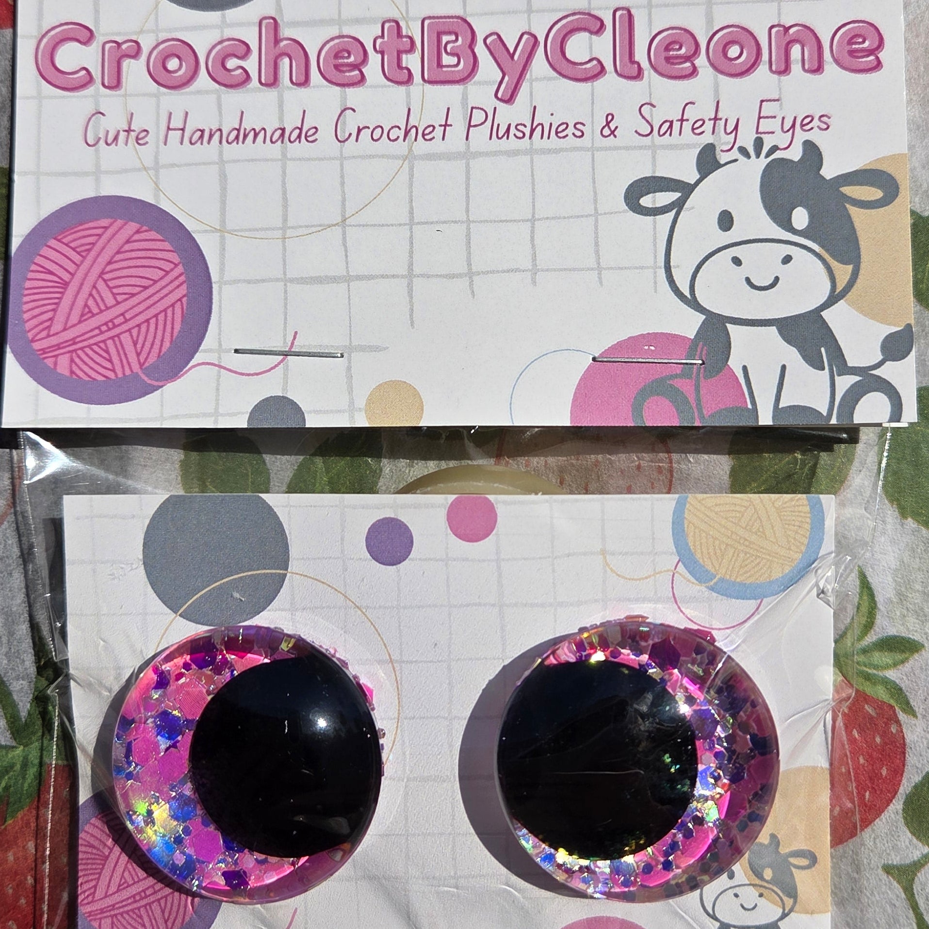Two round black safety eyes with pink and silver glittery cheeks, packaged on a white card labeled 'CrochetByCleo' and placed on a strawberry-patterned fabric surface.