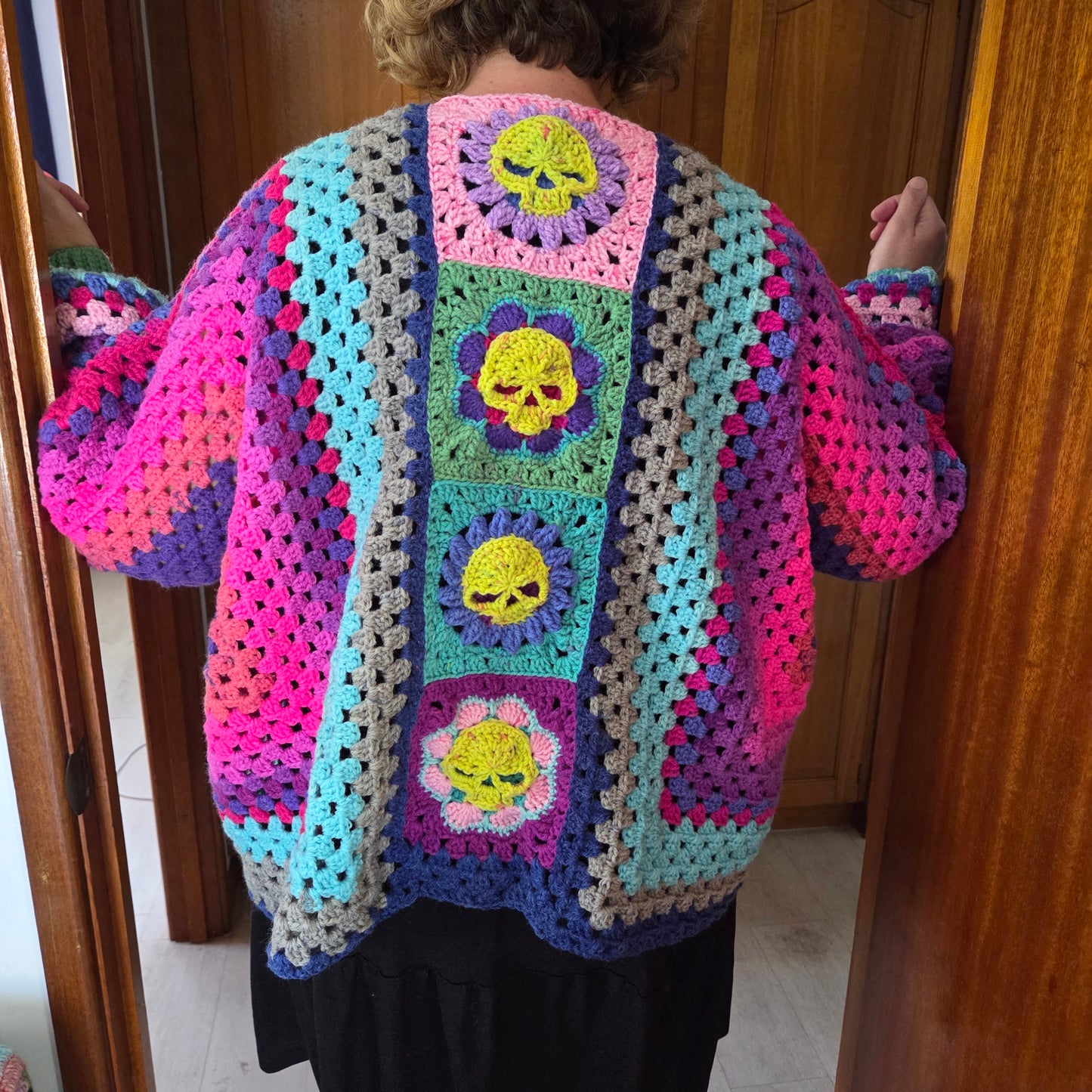 A person viewed from behind wearing a colorful handmade crochet cardigan with floral and skull granny square patterns, standing in a doorway.
