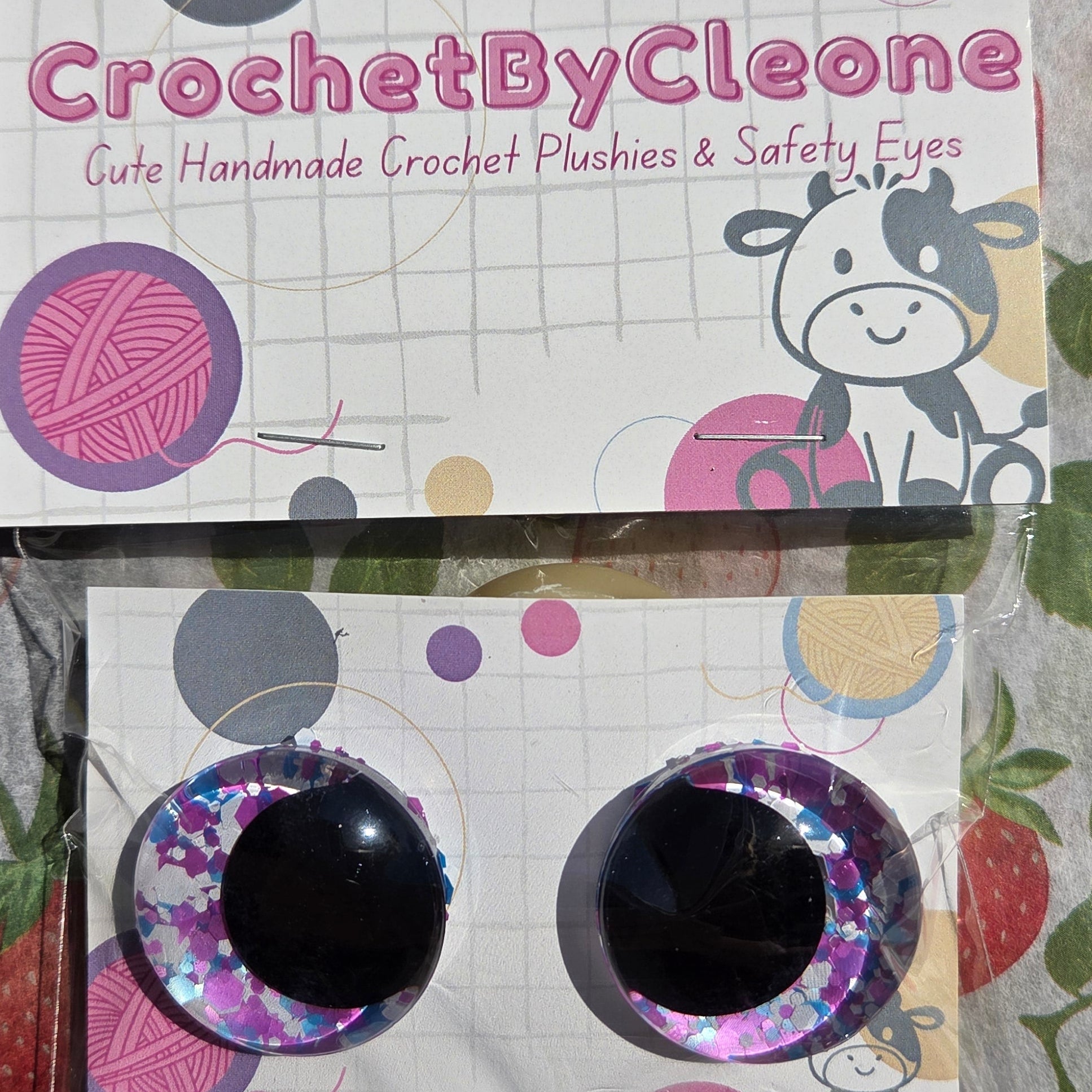 Two round craft eyes with glittery purple and pink rims and black centers, displayed on a printed card labeled 'CrochetByCleone' over a strawberry-patterned fabric background.