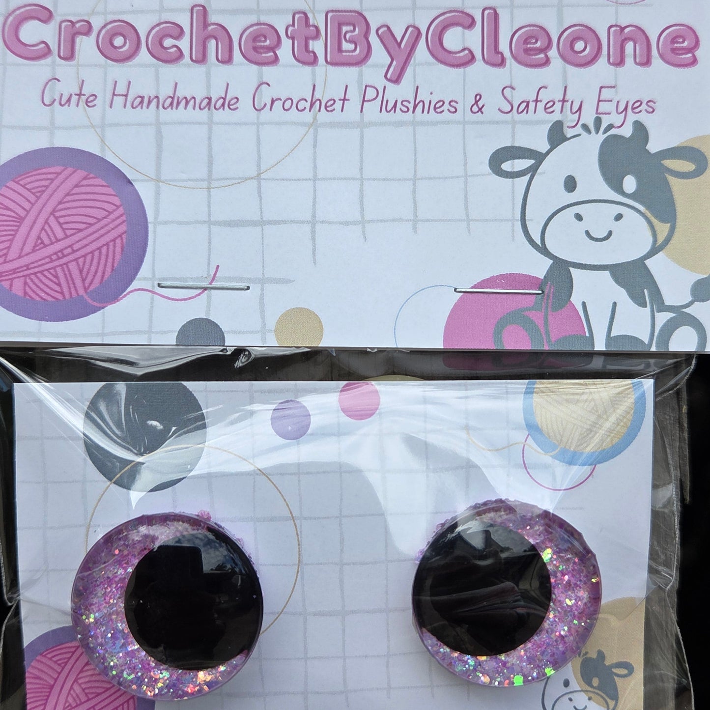 A pair of handmade crochet punch eyes in glittery pink and purple with black centers, displayed in their clear plastic packaging next to the printed brand card 'CrochetByCleo'.