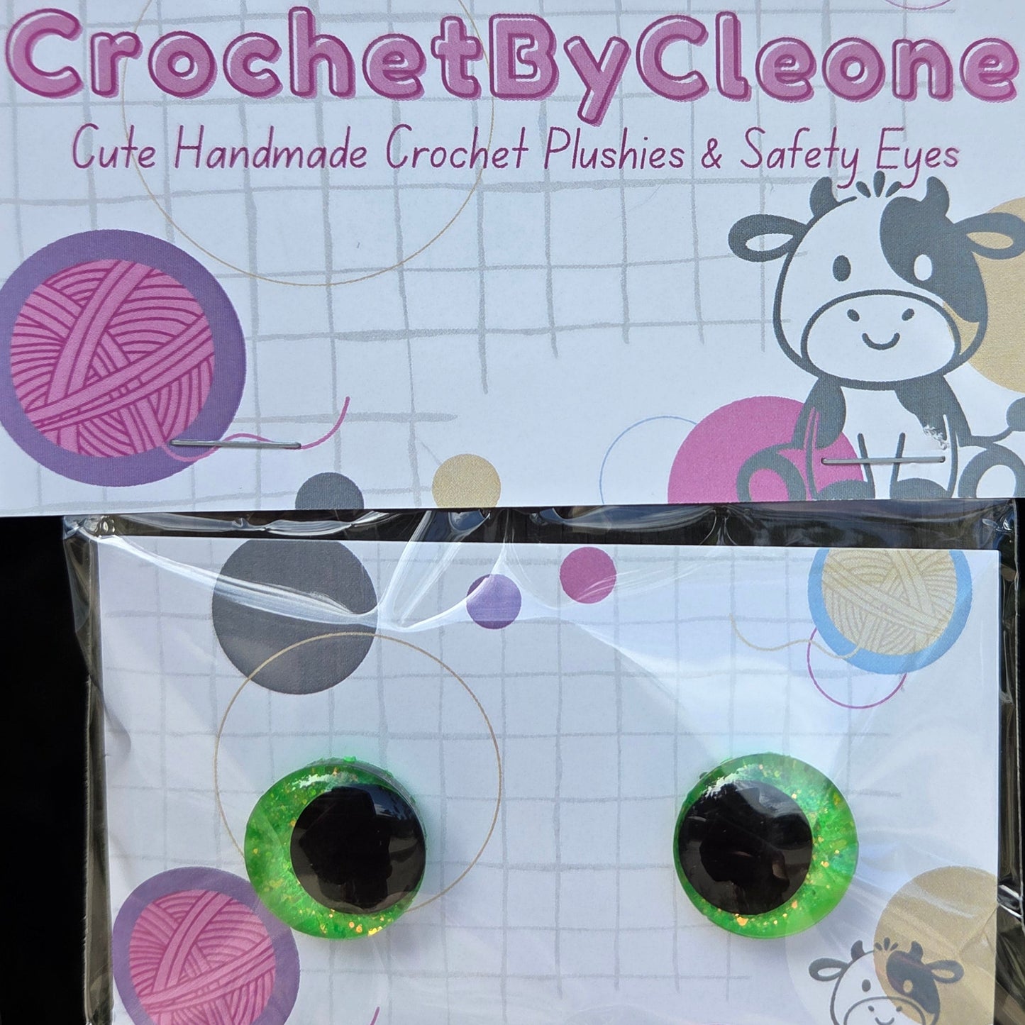 A pair of handmade green glittery safety eyes with black centers, packaged on a branded card that reads 'CrochetByCleone' and says 'Gute Handmade Crochet Plushies & Safety Eyes,' photographed against a rustic wooden background.