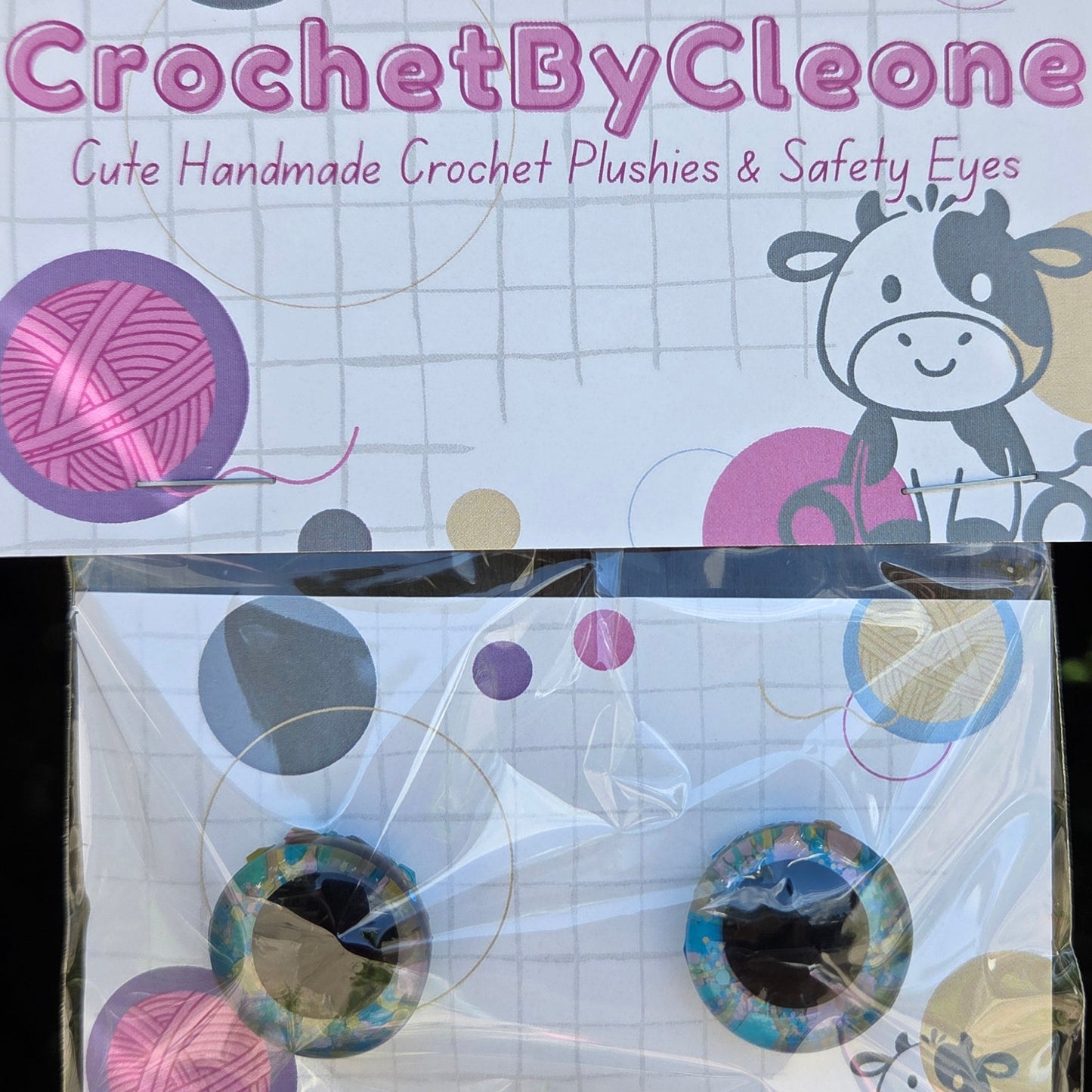 Crochet crochet plushies and safety eyes in a clear plastic bag on a printed card, displayed on a rustic wooden surface. The card features the brand name 'CrochetByCleone' and illustrations of a cow and yarn balls.