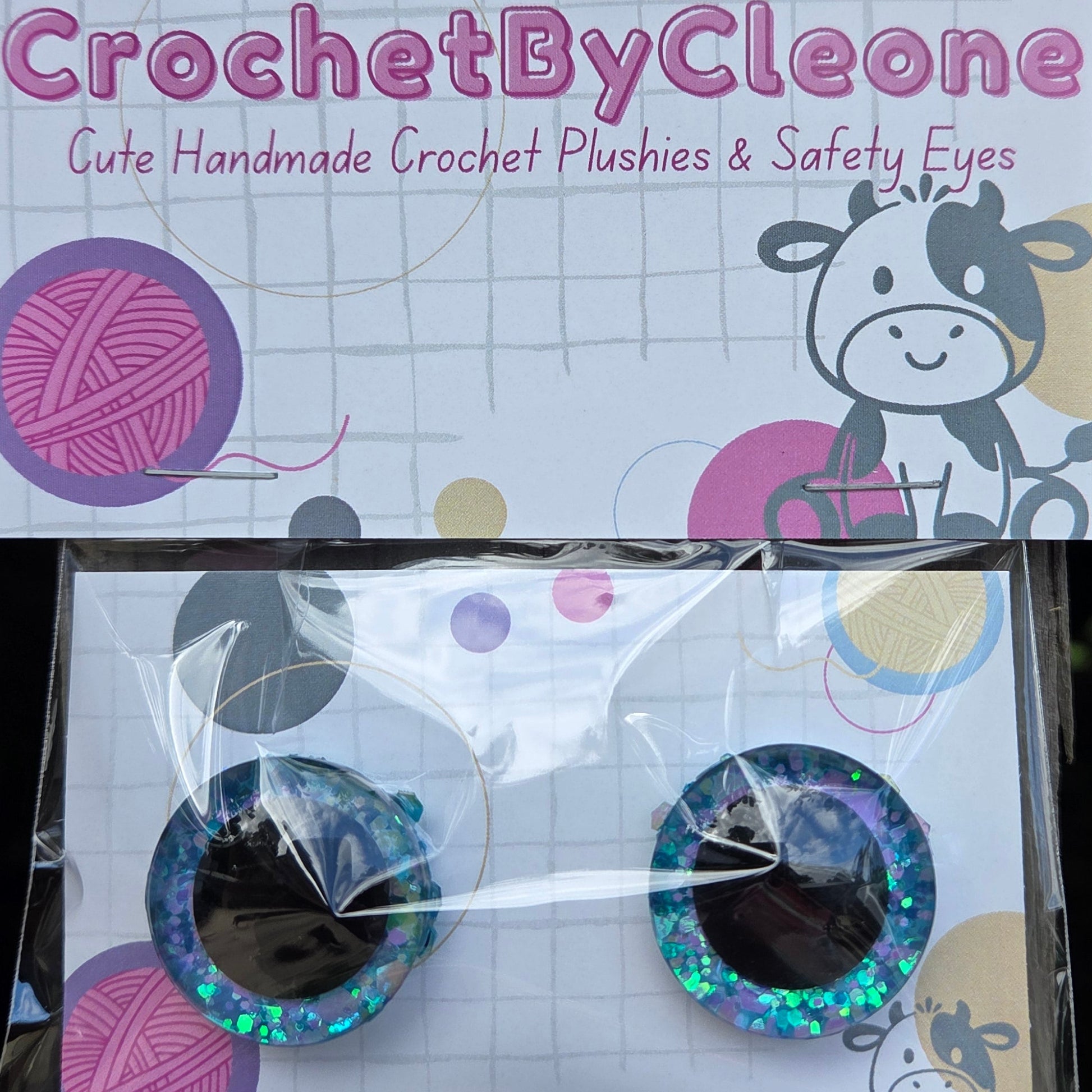 A pair of handmade crochet crochet ears and safety eyes for pets, shown in their packaging. The ears are decorated with iridescent blue glitter and black centers, attached to a white card with pink and purple graphics.