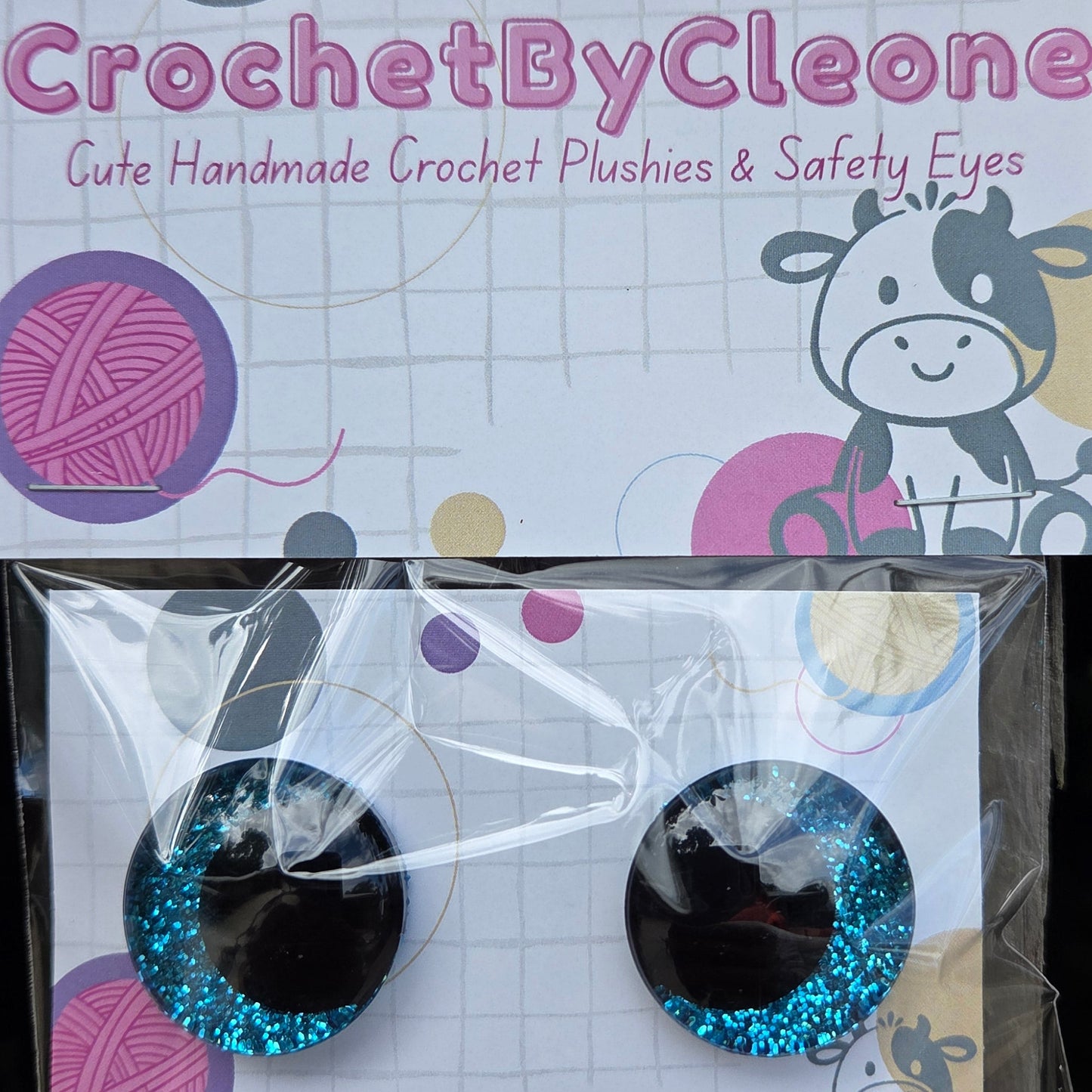 A pair of handmade crochet safety eyes with glittery blue edges and black centers, packaged in a clear plastic sleeve on a printed backing card. The card features the brand name 'CrochetByCleone' and a cartoon cow illustration, set against a rustic wooden background.