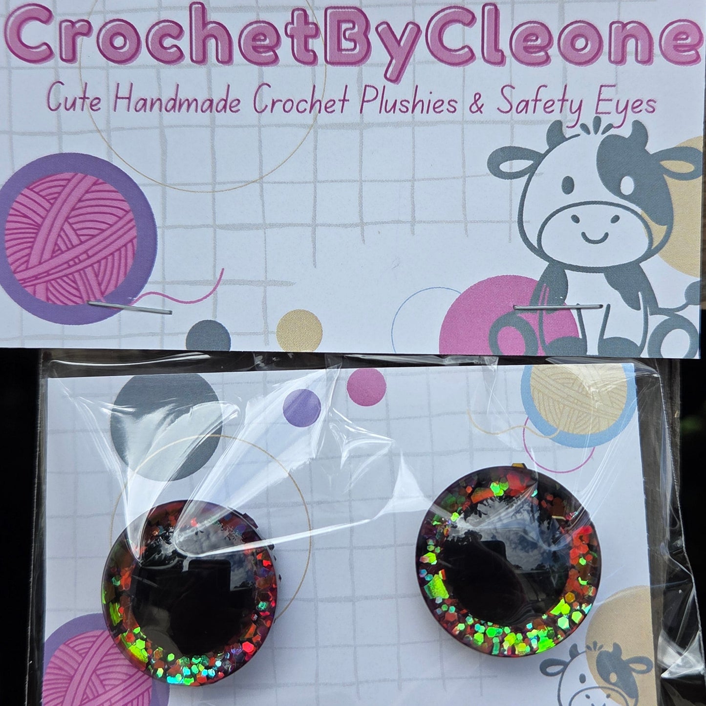 A pair of handmade crochet safety eyes with black centers and colorful glittery edges, displayed on a branded backing card next to its packaging on a rustic wooden surface.