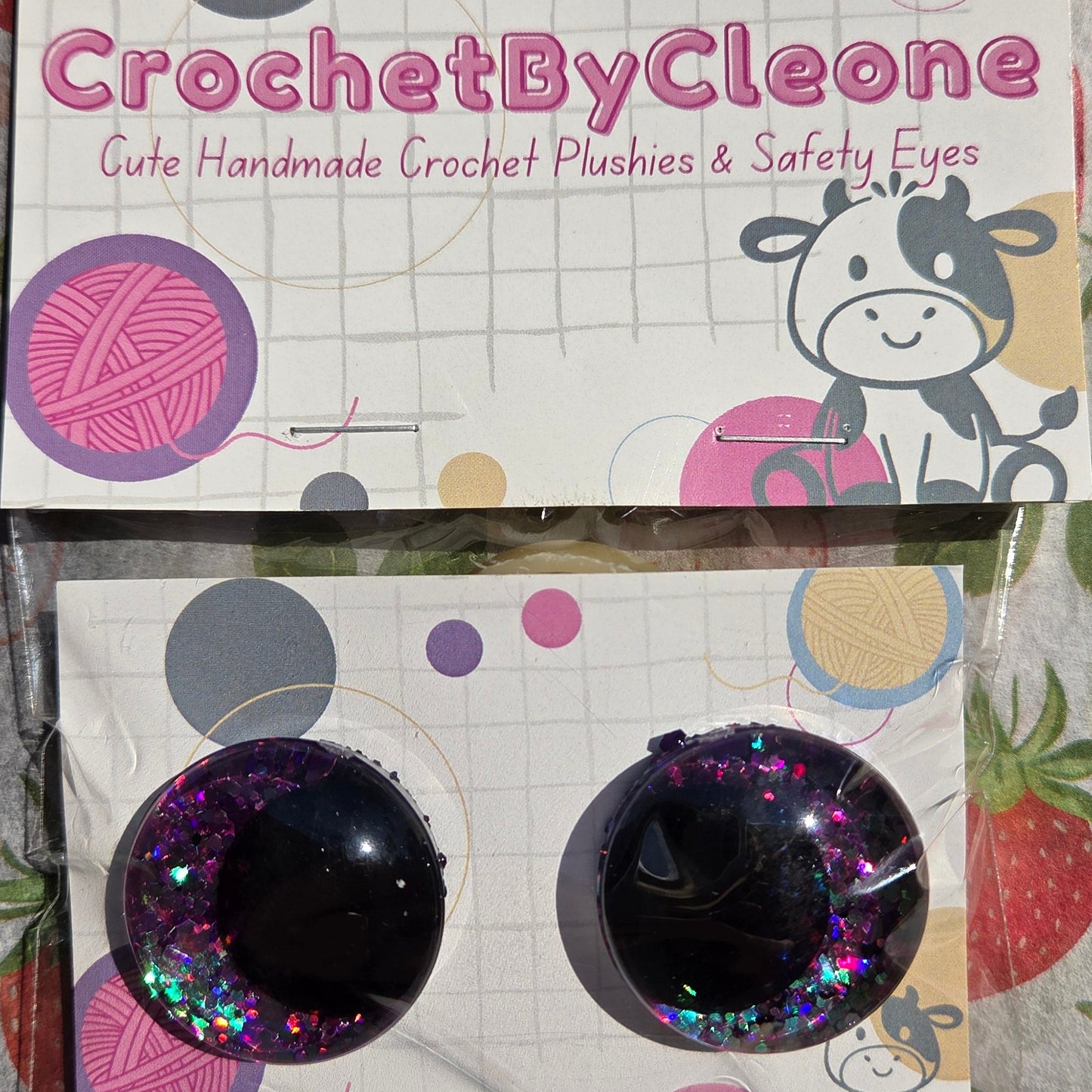A pair of handmade crochet push-on eyes in a clear plastic sleeve, displayed next to their branded packaging card on a strawberry-patterned fabric background. The eyes are black with holographic glitter.