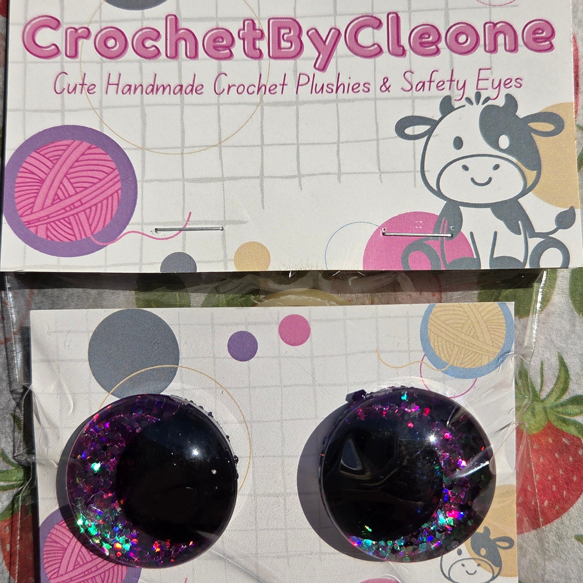 A pair of handmade crochet push-on eyes in a clear plastic sleeve, displayed next to their branded packaging card on a strawberry-patterned fabric background. The eyes are black with holographic glitter.