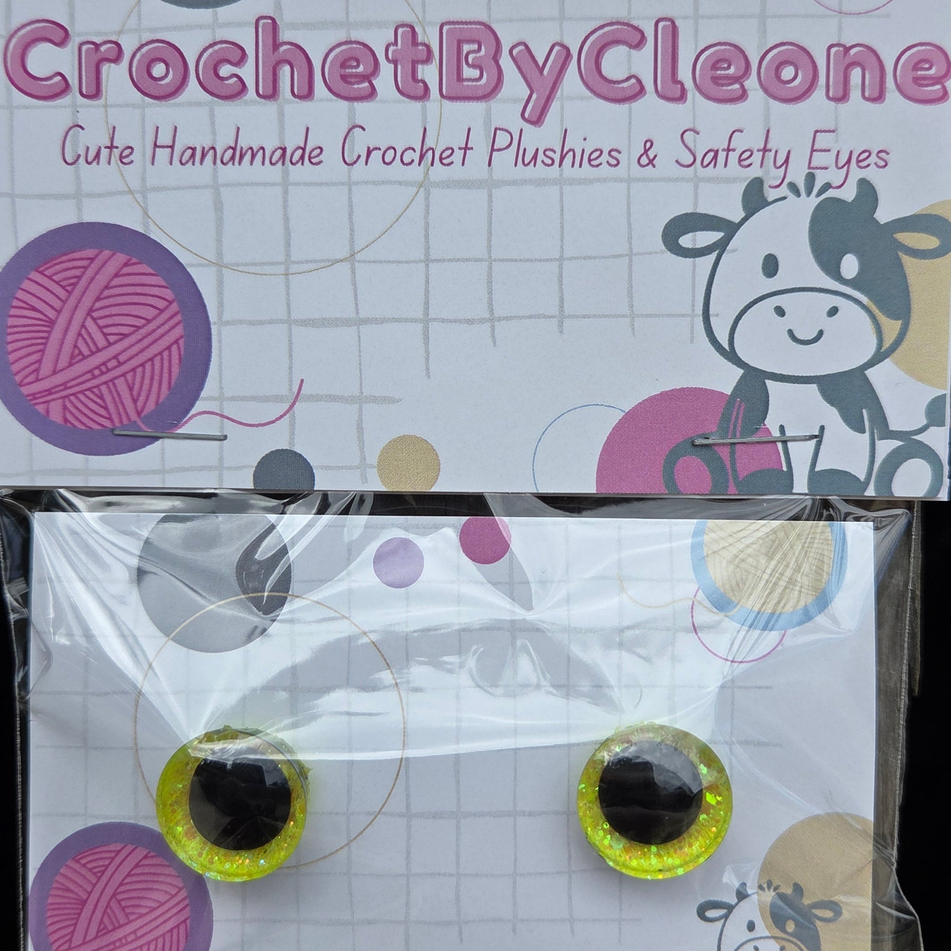 A pair of handmade crochet plush eyes in their retail packaging, displayed on a rustic wooden surface. The packaging features a white grid background with colorful yarn ball graphics, a cow illustration, and the brand name 'CrochetByCleo' printed vertically.
