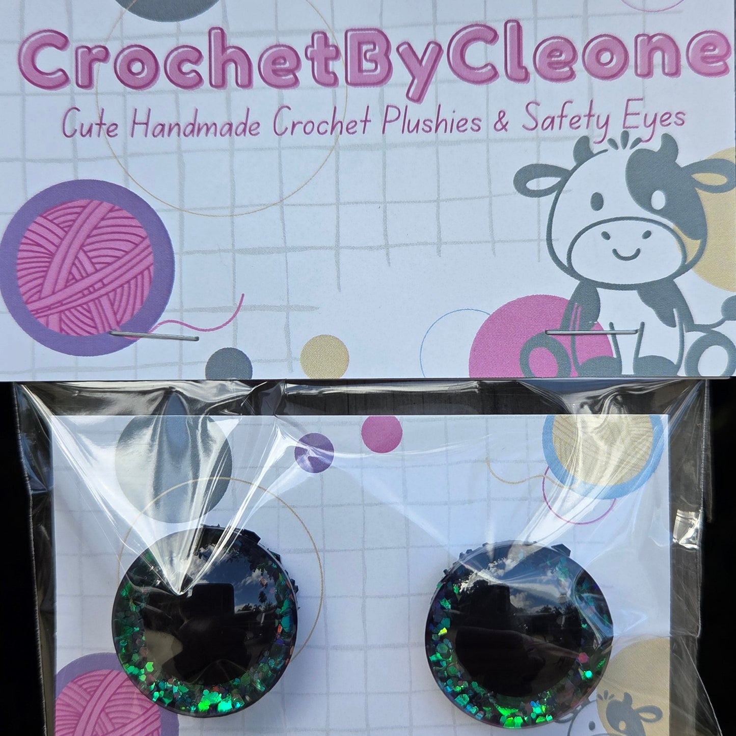 A pair of handmade crochet punch eyes in a clear plastic bag with a branded card, next to the card itself. The punch eyes are black with iridescent green glitter.