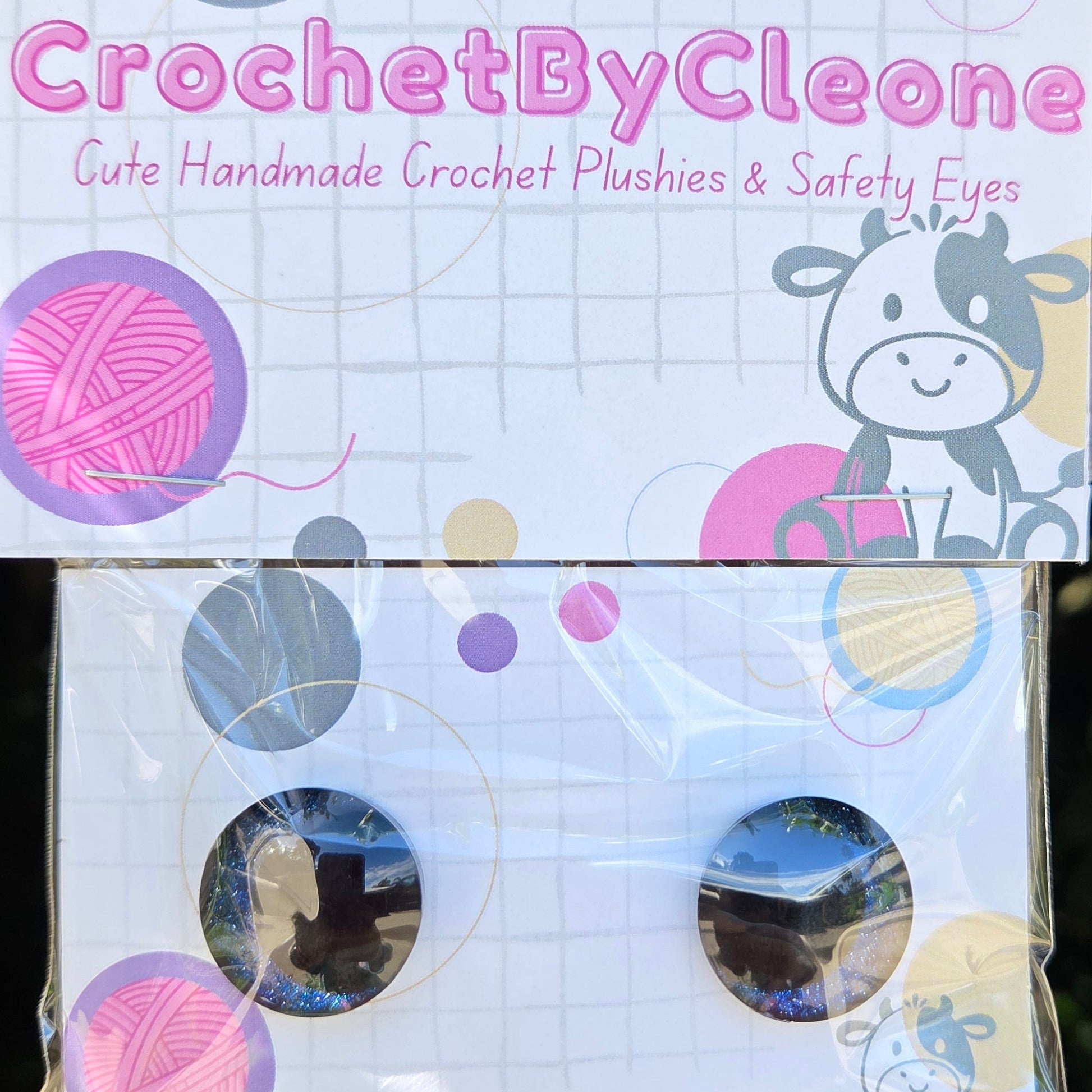 A pair of handmade crochet safety eyes in their original packaging, displayed on a rustic wooden surface. The packaging features a white background decorated with pink yarn balls, purple dots, and a cartoon cow illustration.